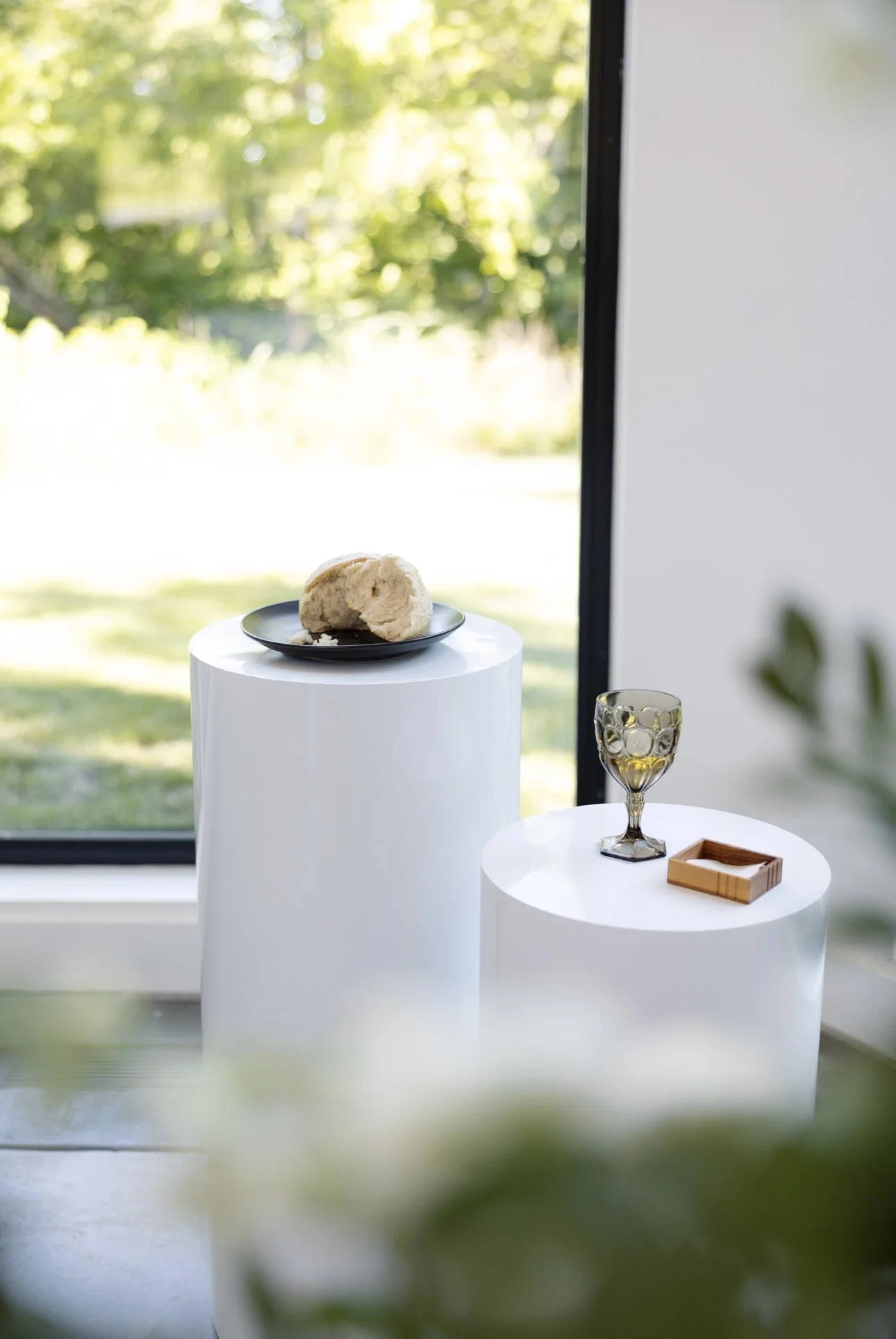 Two white cylindrical pedestals with minimalist decorative objects; one with a black plate holding a piece of bread, the other with a glass and a small wooden tray, set near a large window with green trees outside.