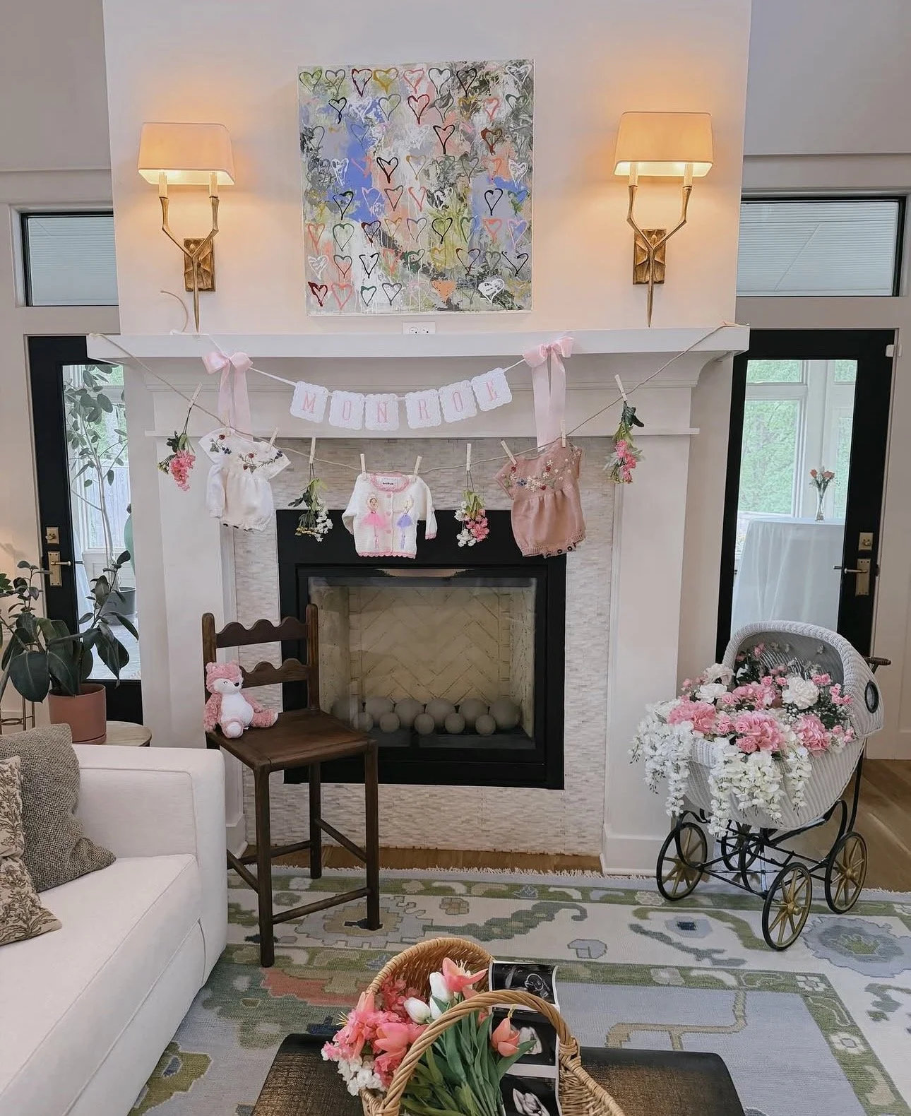 Living room decorated for a celebration with a curved mantel, pink and white flowers, plush toys, and a banner reading 'Monroe' with pink bows, floral art above the mantel, and cozy furniture.