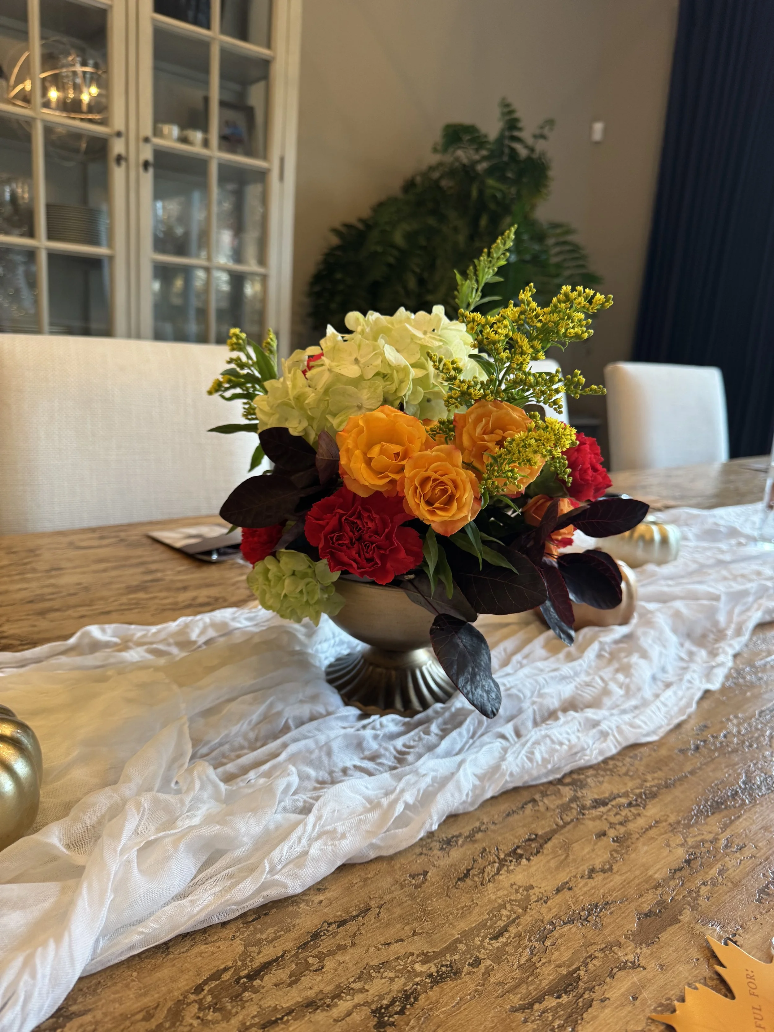 A floral arrangement with white, orange, yellow, red, and green flowers on a beige table runner in a dining room.