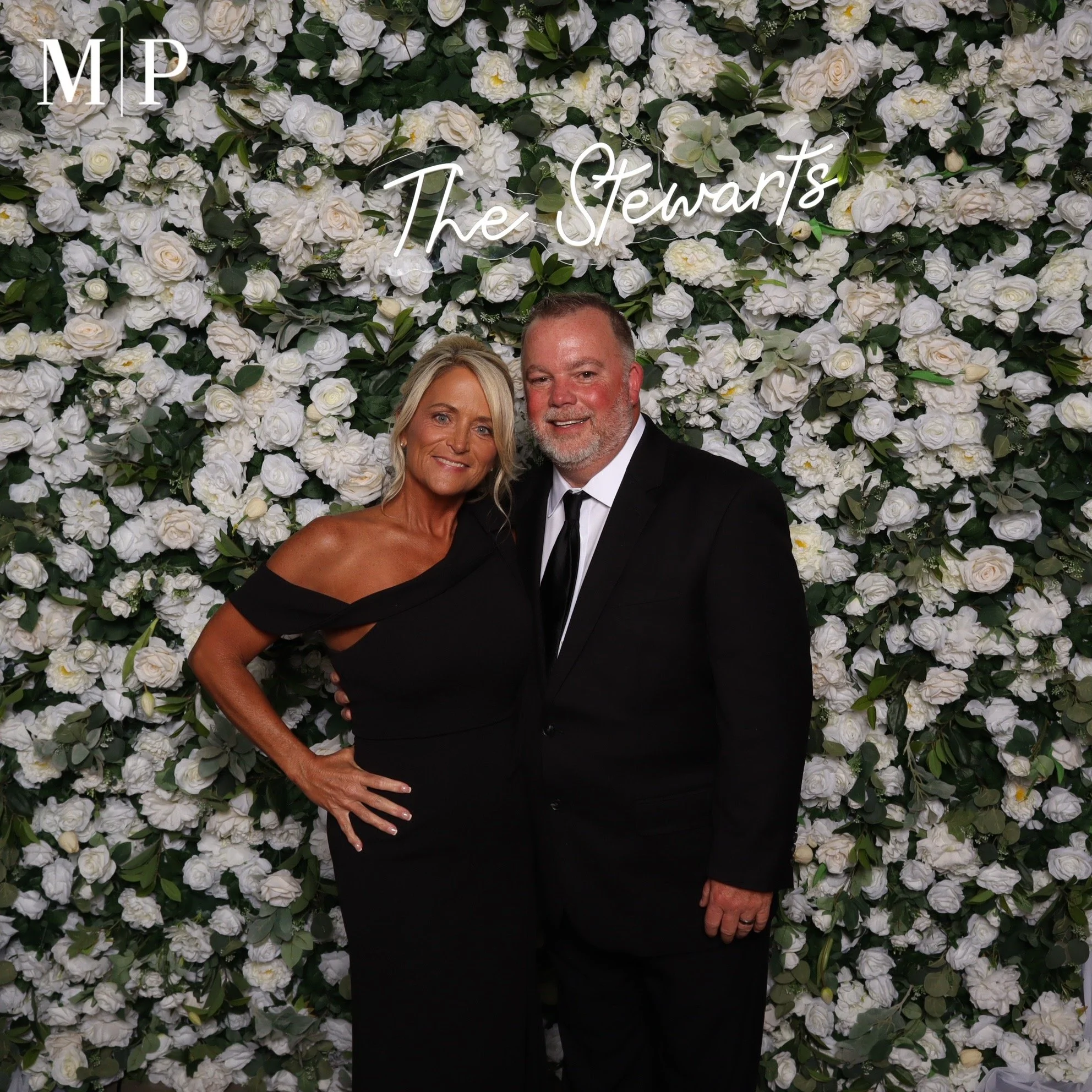 A couple dressed in formal attire, smiling, standing in front of a large floral wall decorated with white flowers and green leaves, with the words 'The Stewarts' written in white neon lights.