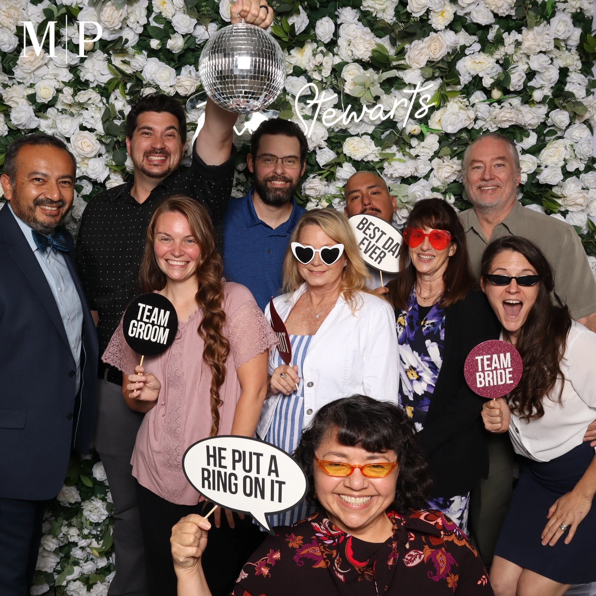 Group of people at a wedding or celebration, posing in front of a white floral backdrop, holding signs and accessories, with a disco ball above them.