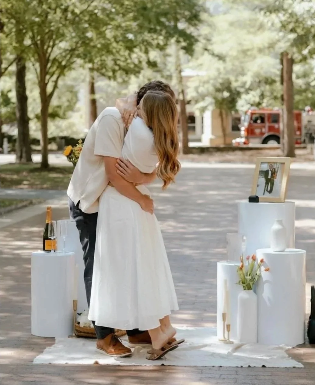A couple embracing at an outdoor wedding ceremony with trees and a fire truck in the background, decorated with vases and a photograph on a white stand.