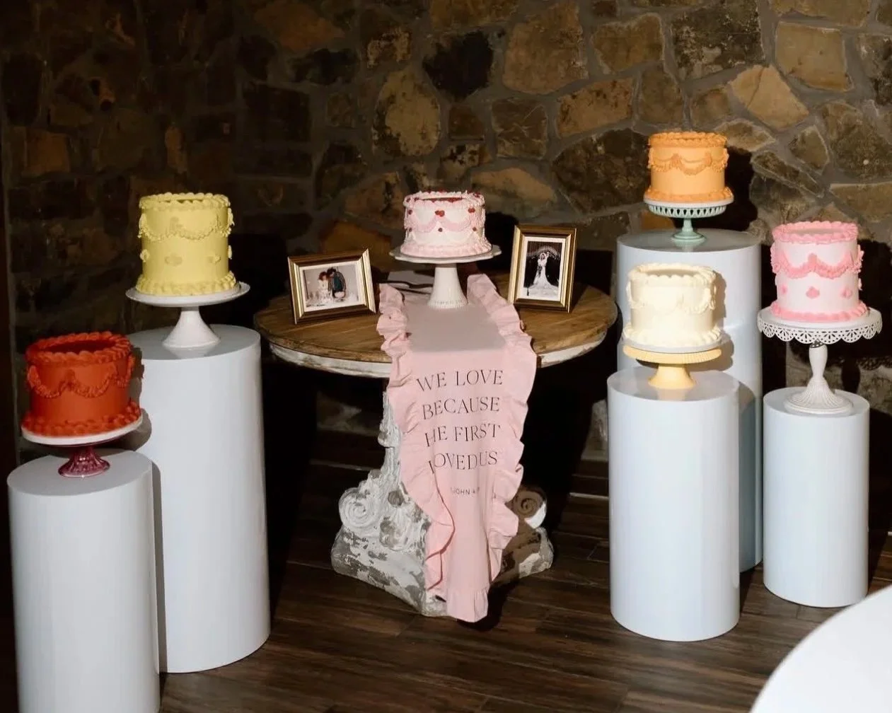 Six decorated wedding cakes displayed on white pedestals around a wooden table with framed wedding photos and a pink fabric banner that reads "WE LOVE BECAUSE HE FIRST LOVED US." The cakes are in yellow, pink, orange, white, cream, and peach colors, against a rustic stone wall background.