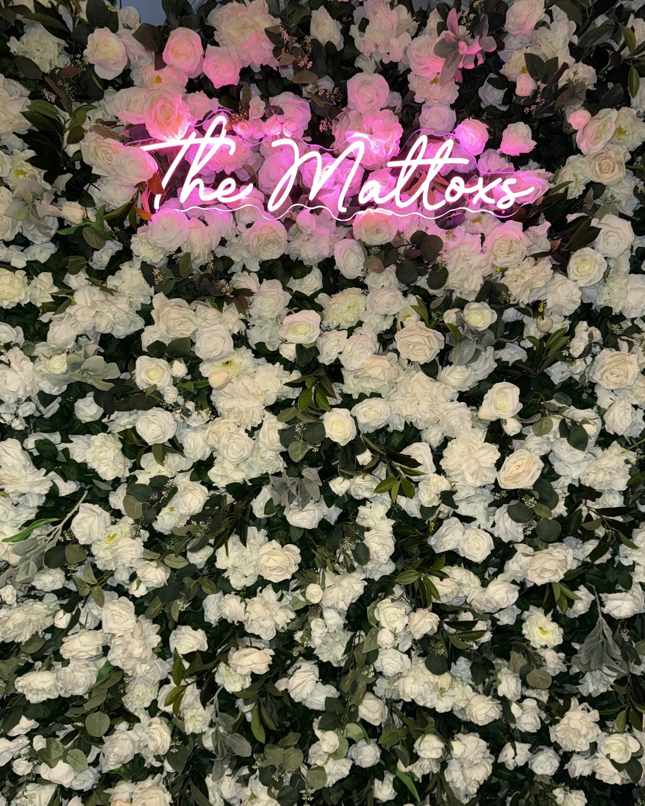 A wall covered with white and light pink roses and green leaves, with a neon pink sign that says 'the Mattox' in cursive.