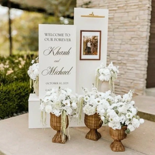 Wedding welcome sign with white floral arrangements and framed photo of the couple, featuring text with names Khanh and Michael, date October 10, 2025, and the words "Welcome to our forever."