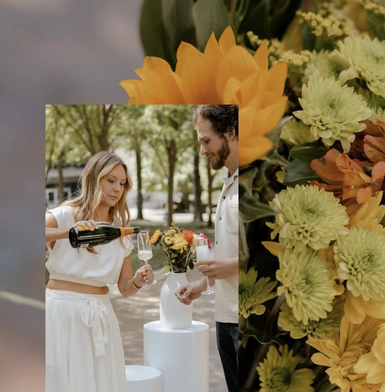 A woman pouring champagne into a glass for a man at a garden celebration with a flower bouquet in a white vase on a white table, with yellow and green flowers and a blurred background of trees.