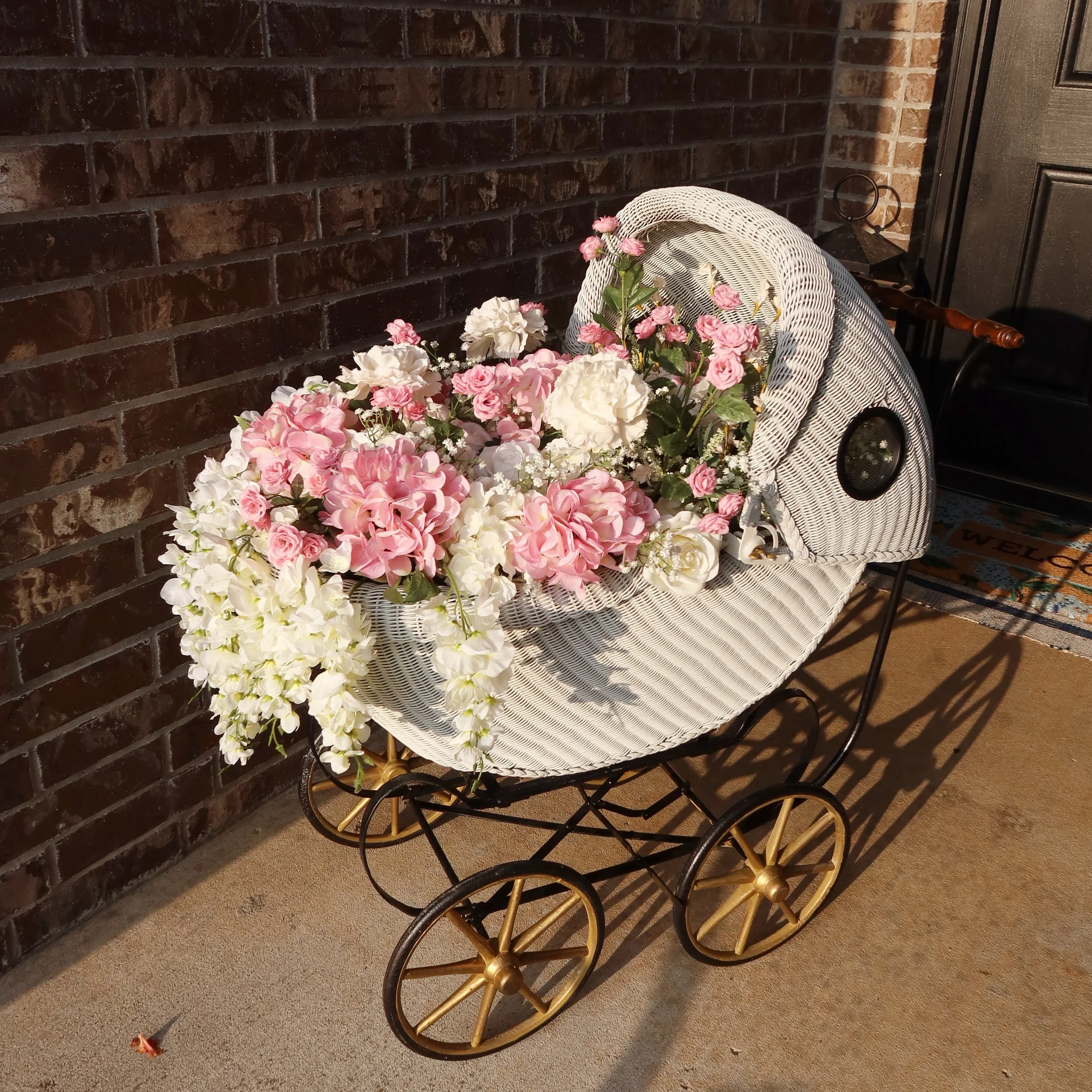 A white wicker baby stroller filled with pink and white artificial flowers parked on a porch next to a brick wall and a black door.