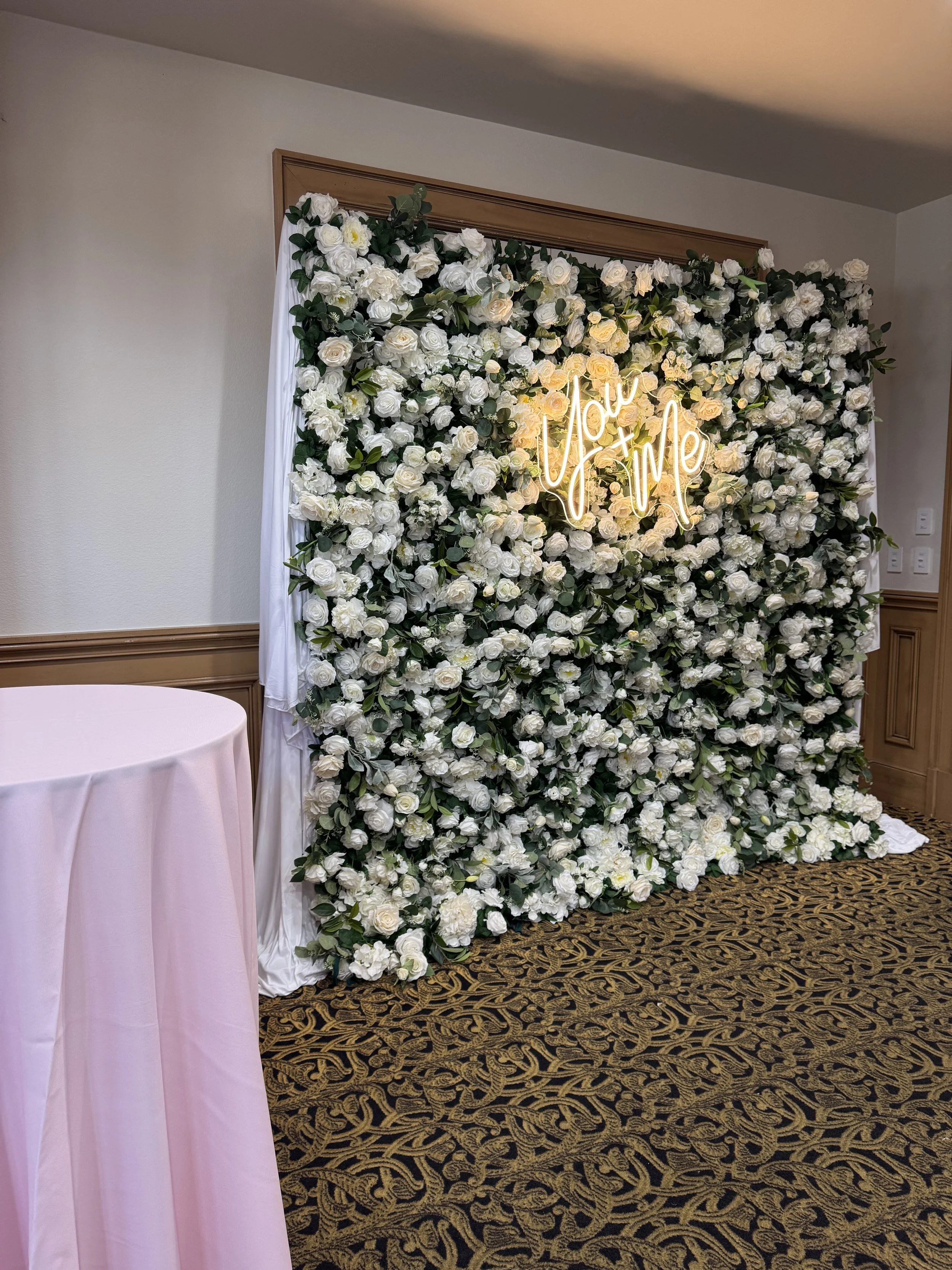 A wall decorated with white and cream roses and green foliage featuring a neon sign that says "Now Wifed" in cursive script.