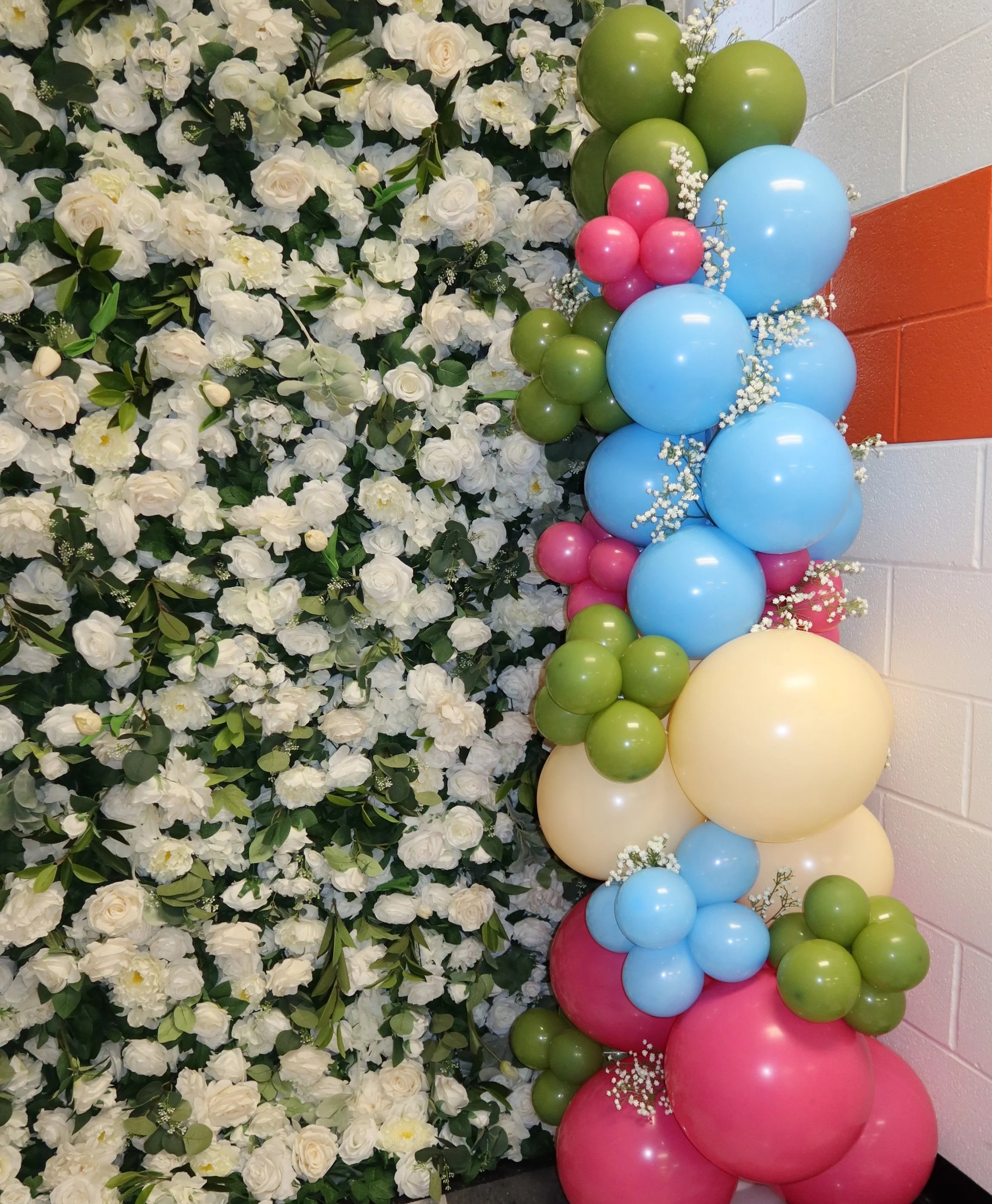 A decorative display of white flowers and green foliage on the left side and a colorful arrangement of balloons in pastel shades of pink, blue, yellow, and green on the right side, against a brick wall background.