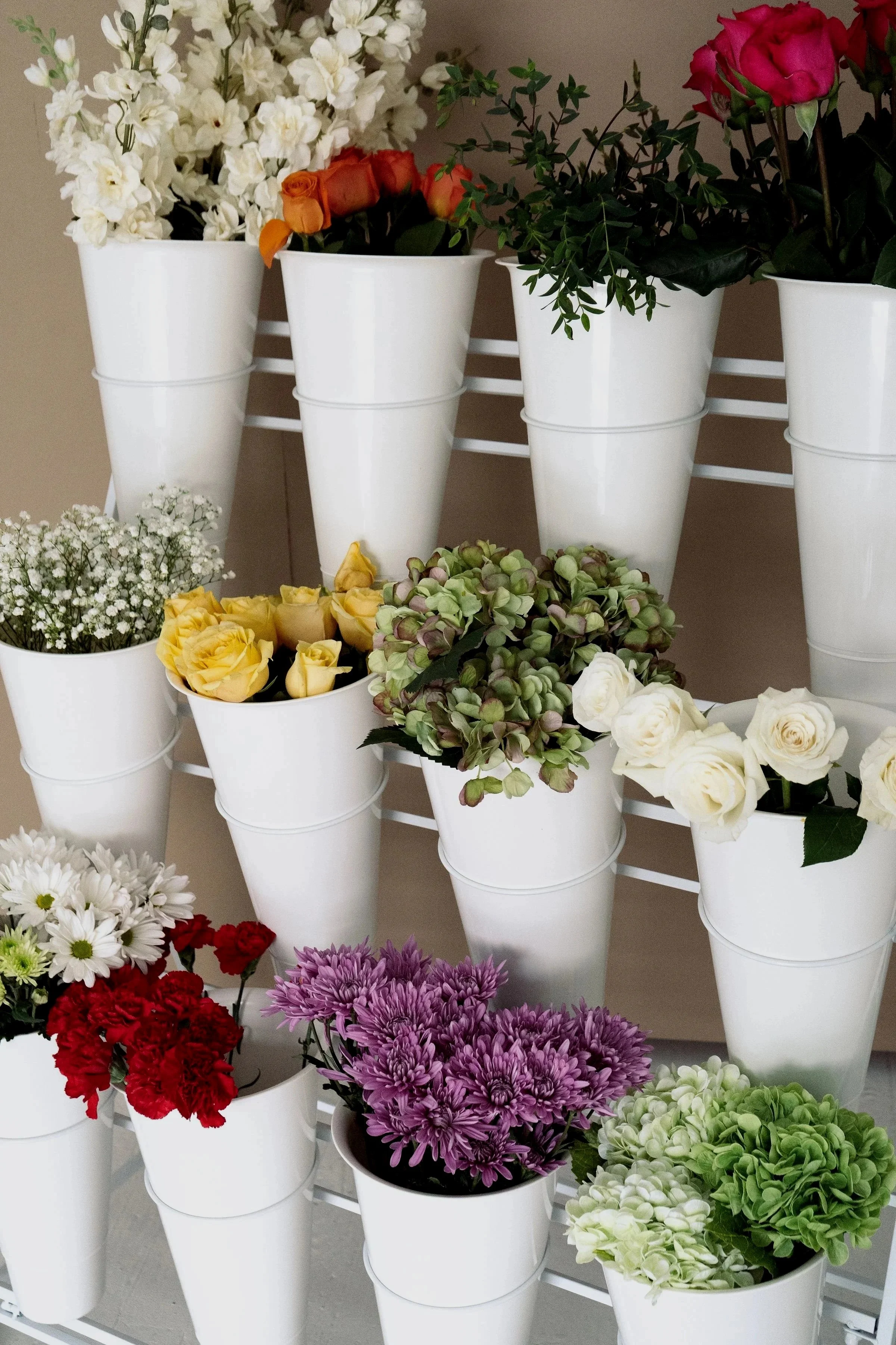 Various flowers in white pots arranged on a metal shelf, including white hydrangeas, pink roses, purple chrysanthemums, yellow roses, and red carnations.