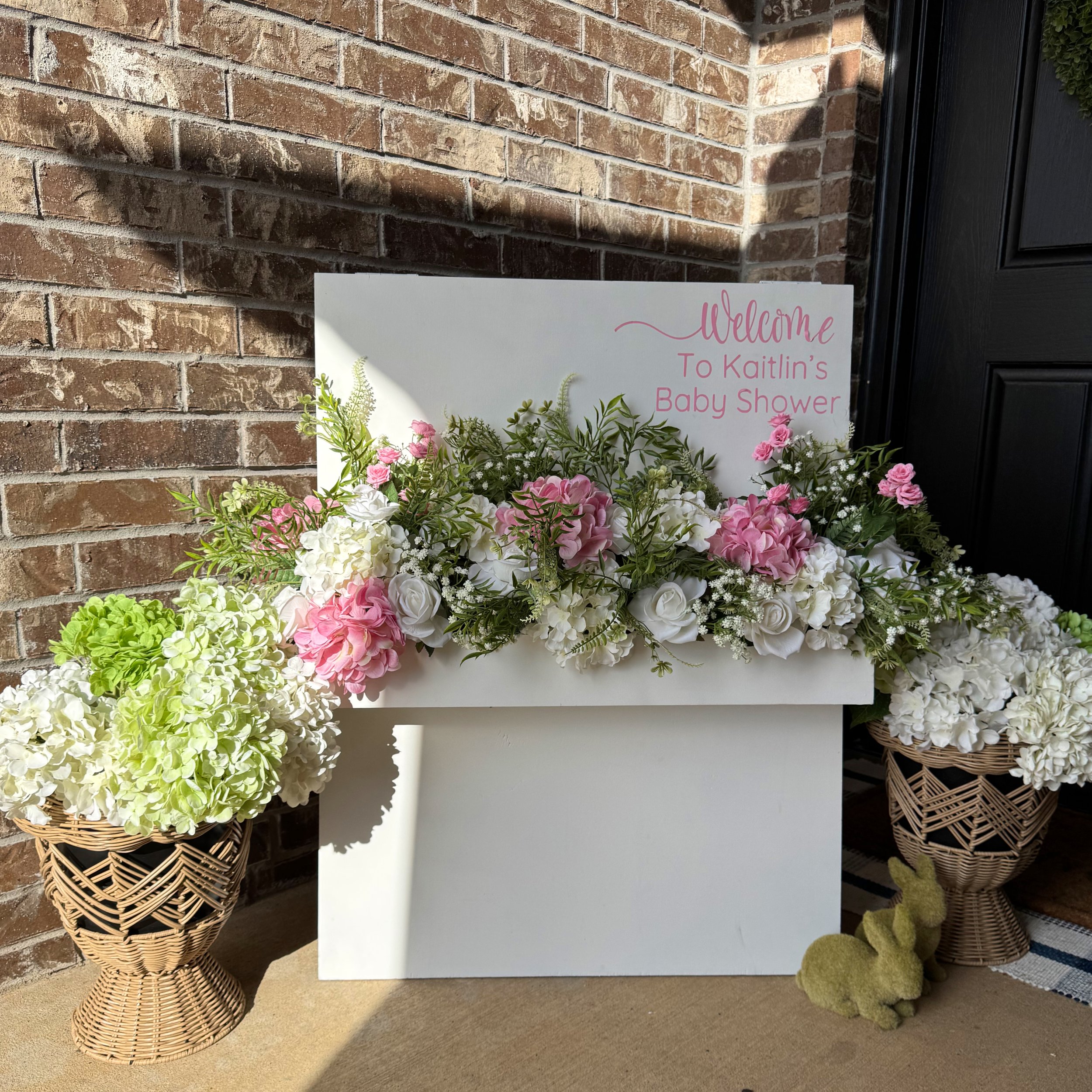 Arrangement of pink and white flowers in baskets with a sign that reads 'Welcome to Kaitlin's Baby Shower' next to a brick wall.