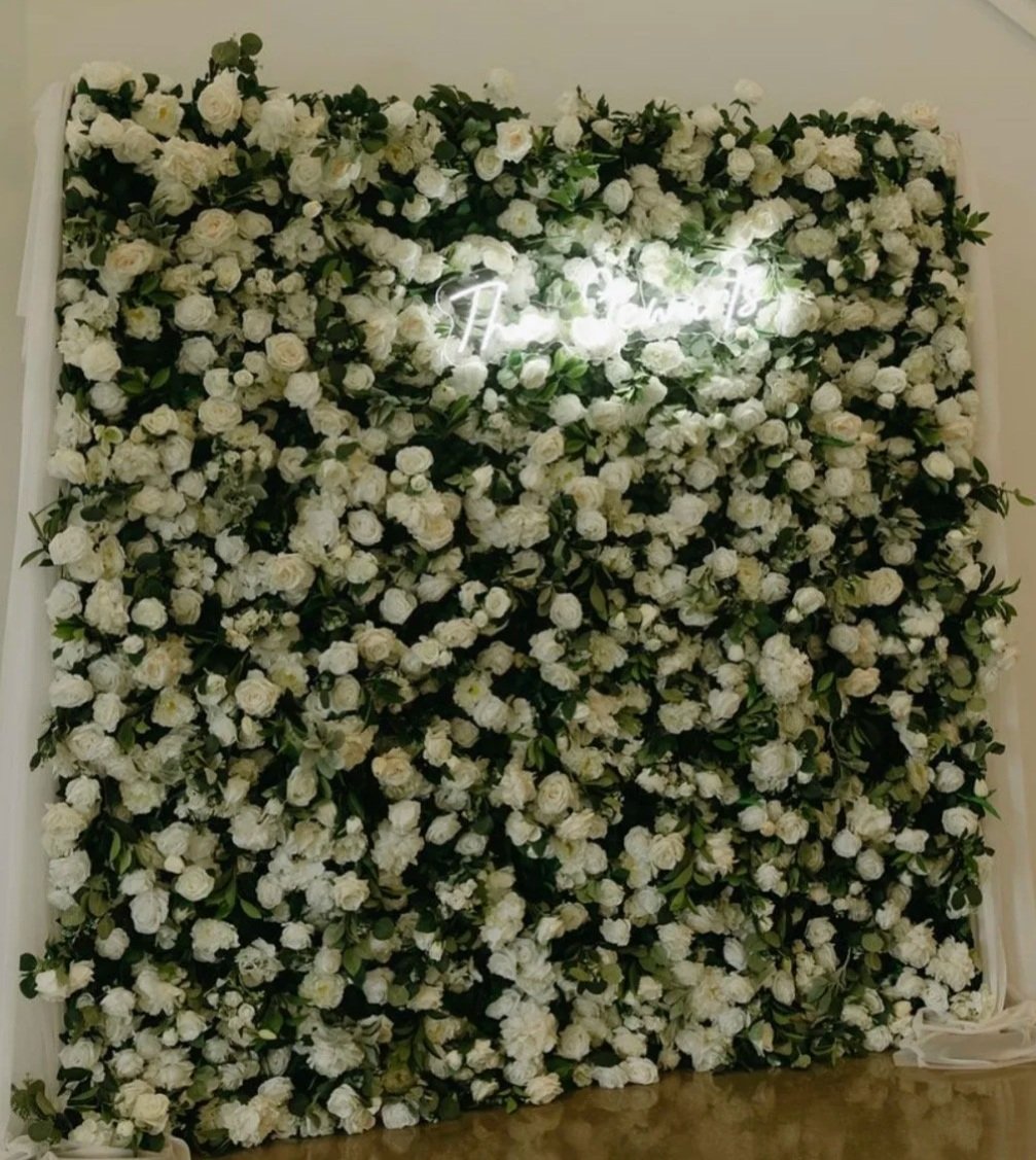 A wall decorated with white flowers and green leaves, with a neon sign in the center that says "The best" in cursive script.