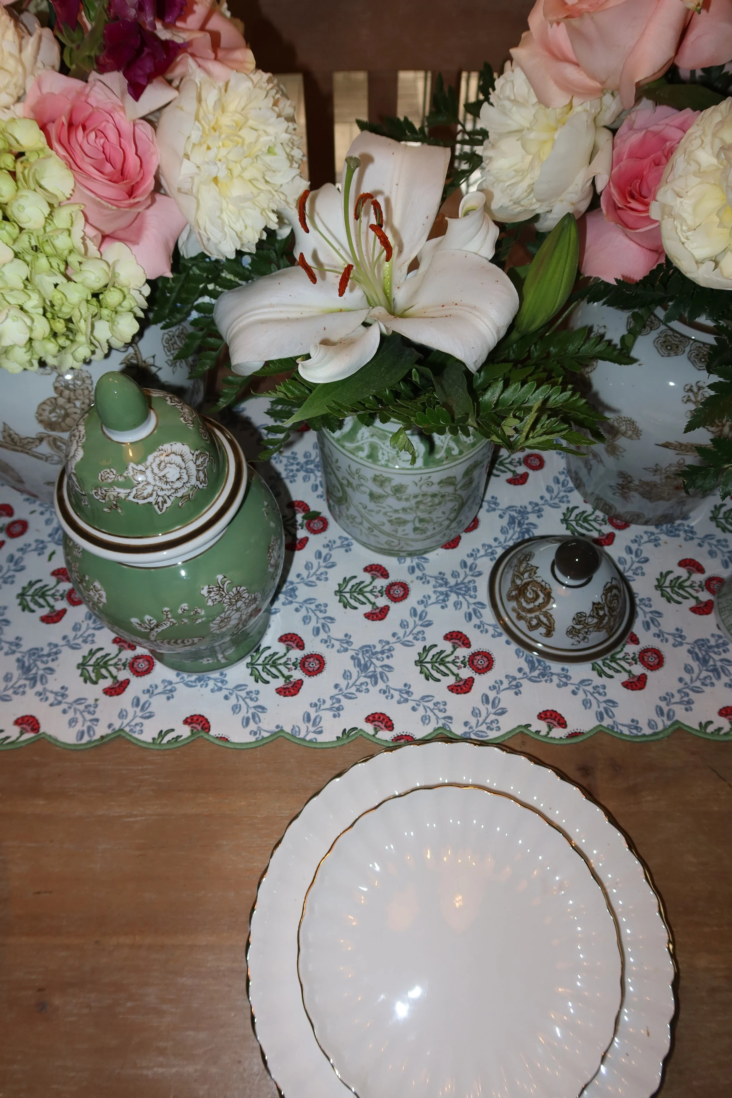 A floral arrangement featuring a white lily, pink roses, white carnations, and green foliage in decorative bowls, placed on a table with a patterned tablecloth. There are also ceramic containers with gold and floral designs on the table.