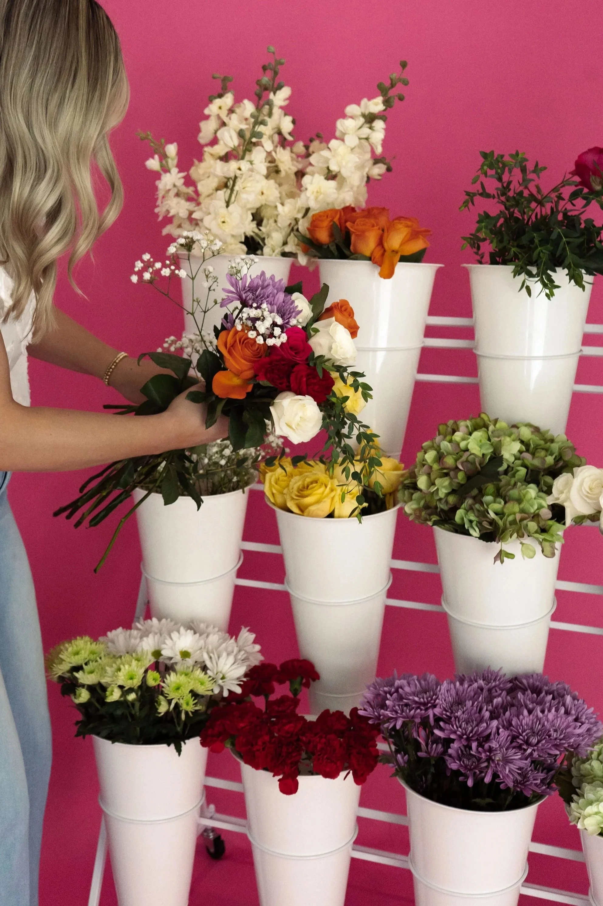 A woman arranging various colorful flowers in white buckets against a pink background.
