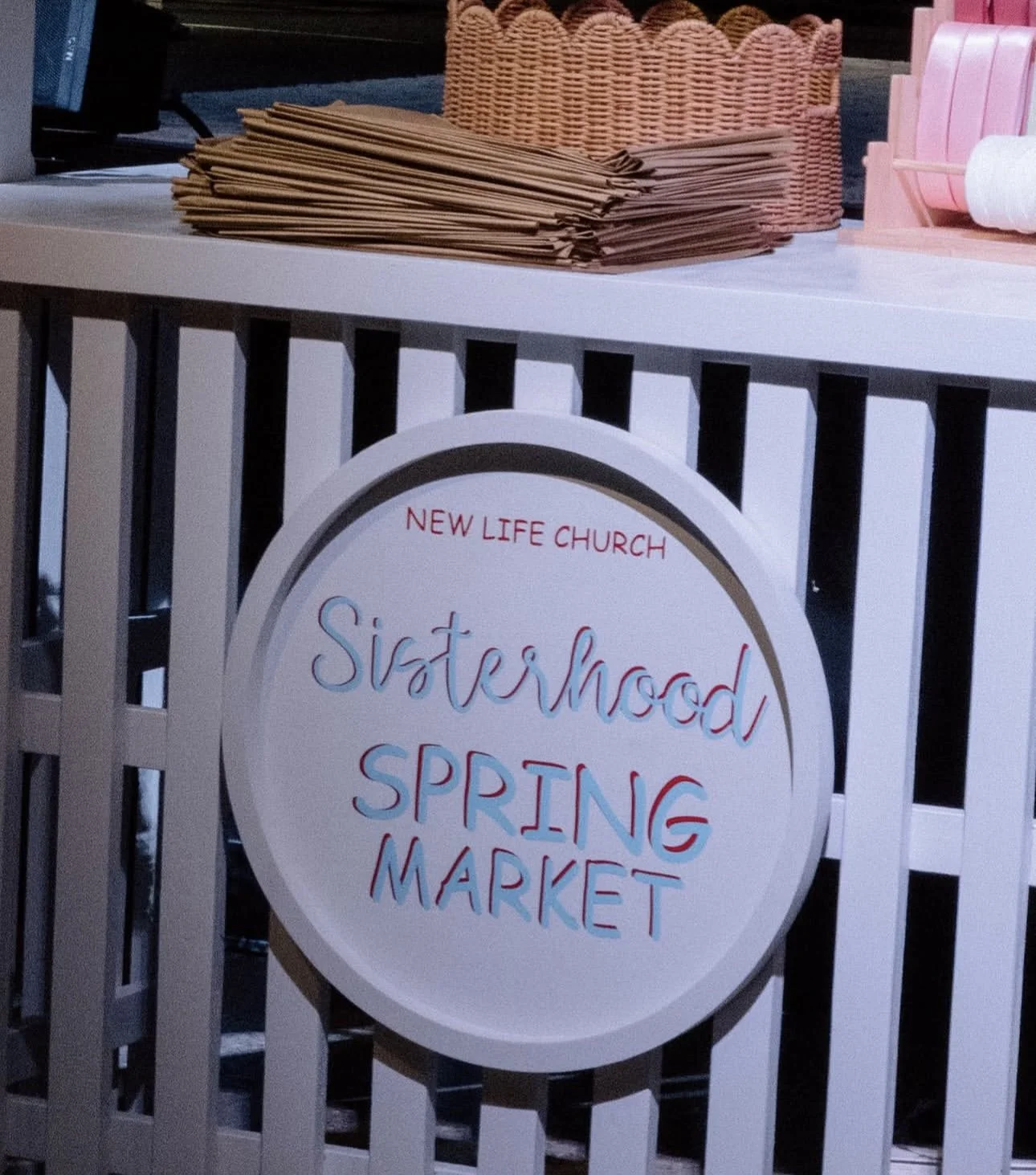 Sign for Sisterhood Spring Market at New Life Church, with brown paper bags and pink baskets on a table behind it.