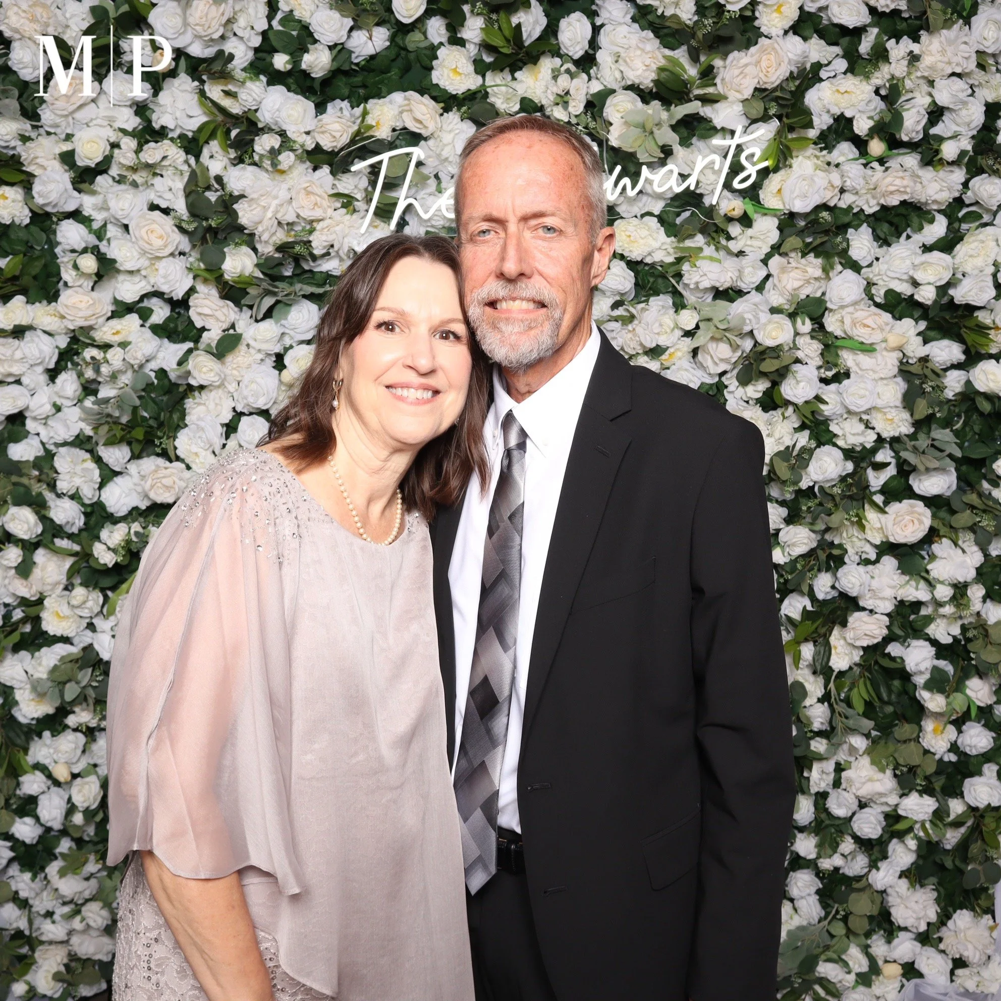 A couple dressed in formal attire standing in front of a wall of white roses at a wedding event.