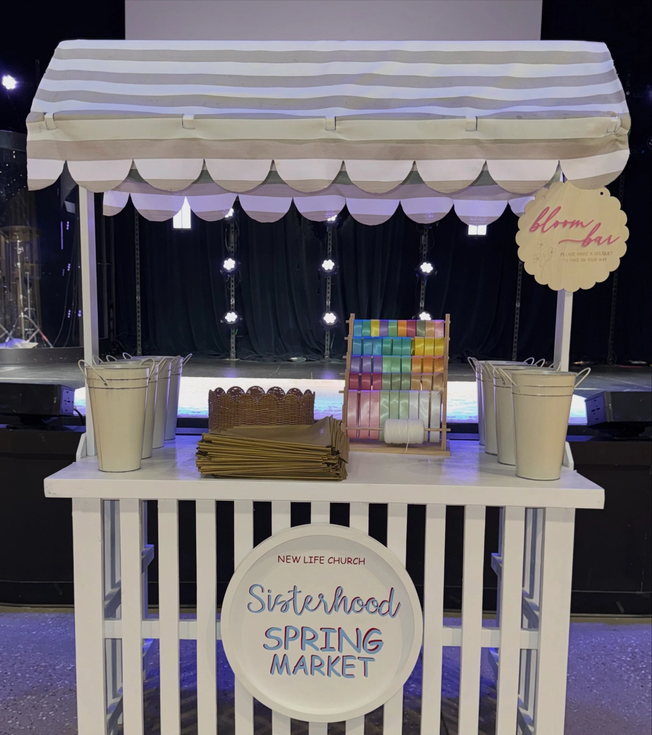 White wooden stand with a sign that reads 'Sisterhood Spring Market' and a smaller sign that says 'New Life Church. Please make a duplicate to take on your way.' Behind the stand, there is a black backdrop with small flower-shaped lights hanging down