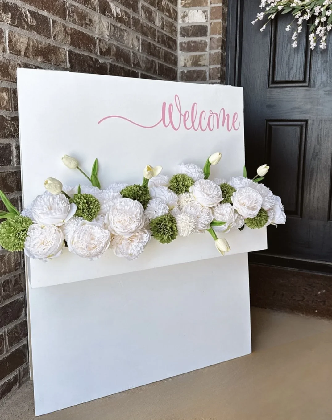 White welcome sign decorated with white and green flowers placed on a porch next to a brick wall and a black door.