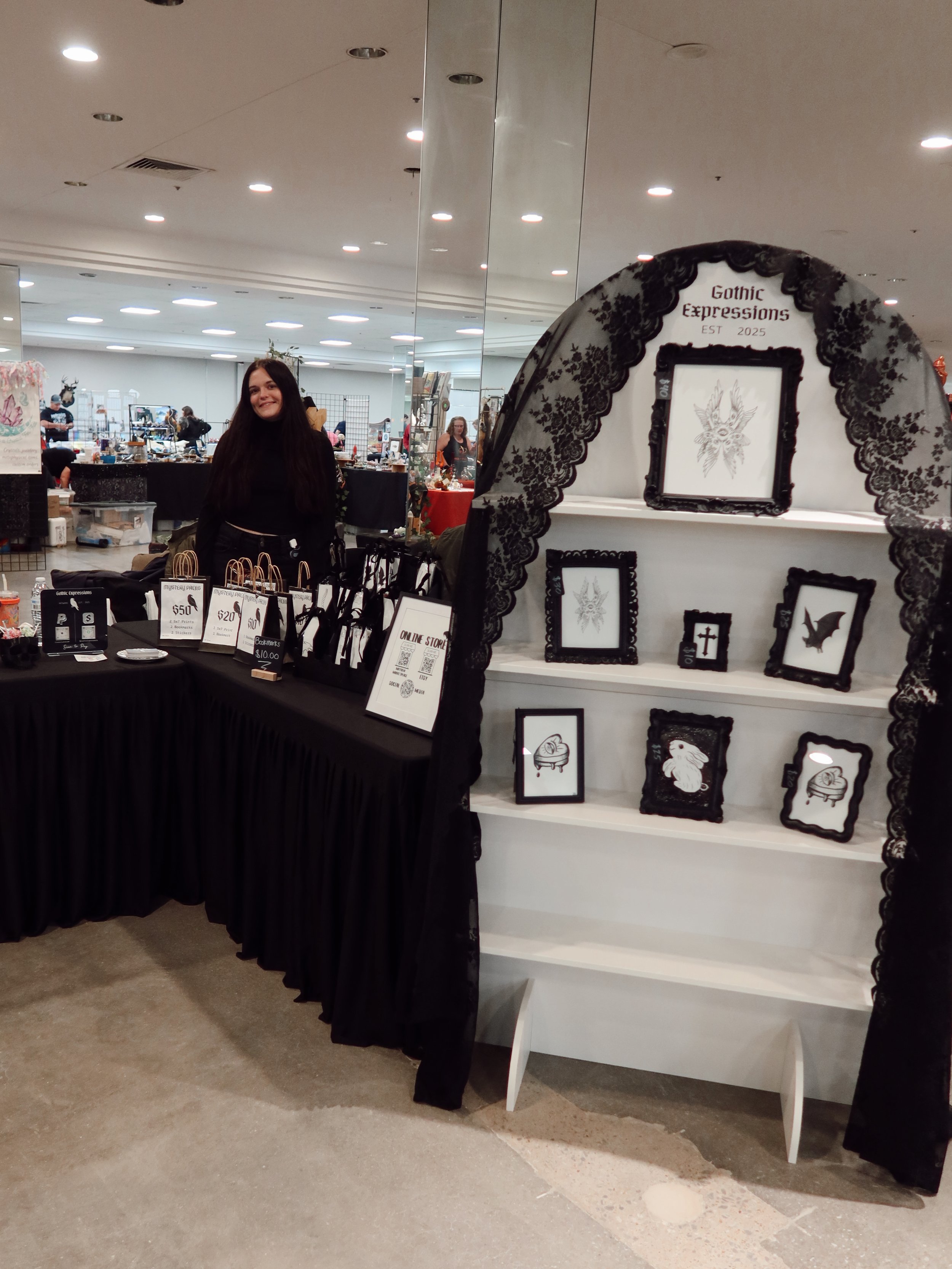 A young woman with long dark hair smiling at a gothic-themed booth with framed artwork, possibly at a convention or fair, with a display wall and other tables in the background.