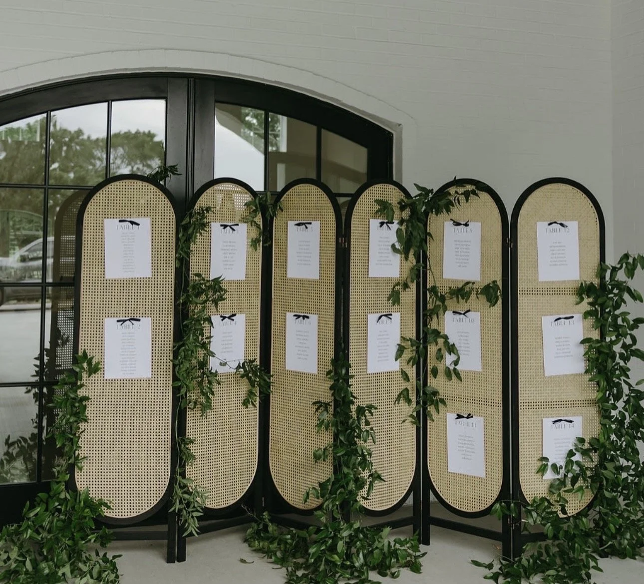 A decorative folding screen with a rattan-like pattern, adorned with green leaves, displaying printed menus on white sheets of paper, placed in a bright room near large windows.