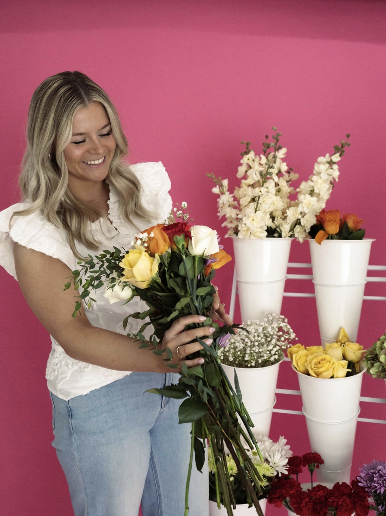 A woman with blonde hair smiling and holding a bouquet of mixed flowers in front of a pink background. There are white pots with various flowers arranged on a white stand behind her.