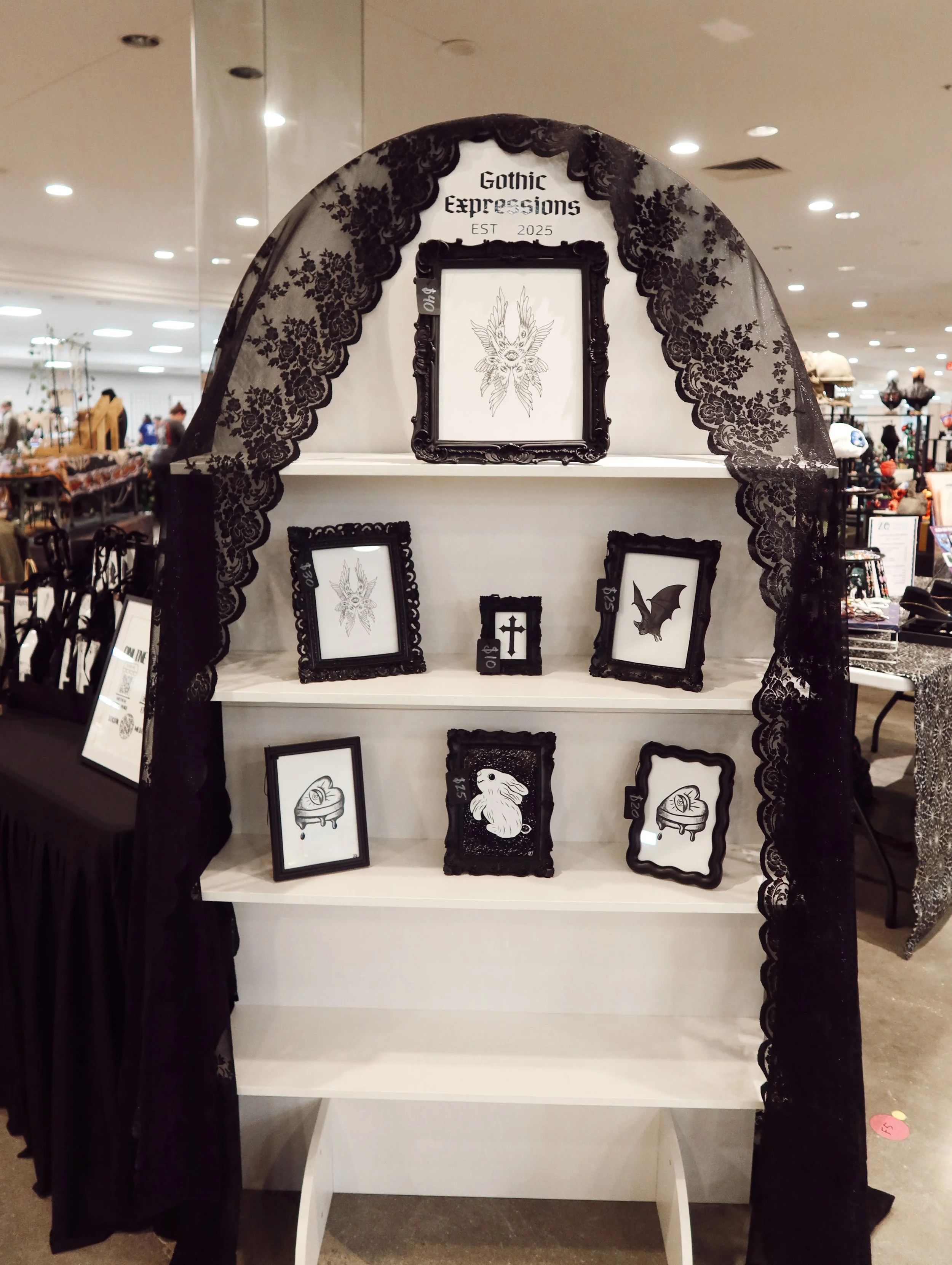Display stand with gothic-themed framed artwork, black lace draped on either side, at a shopping event.