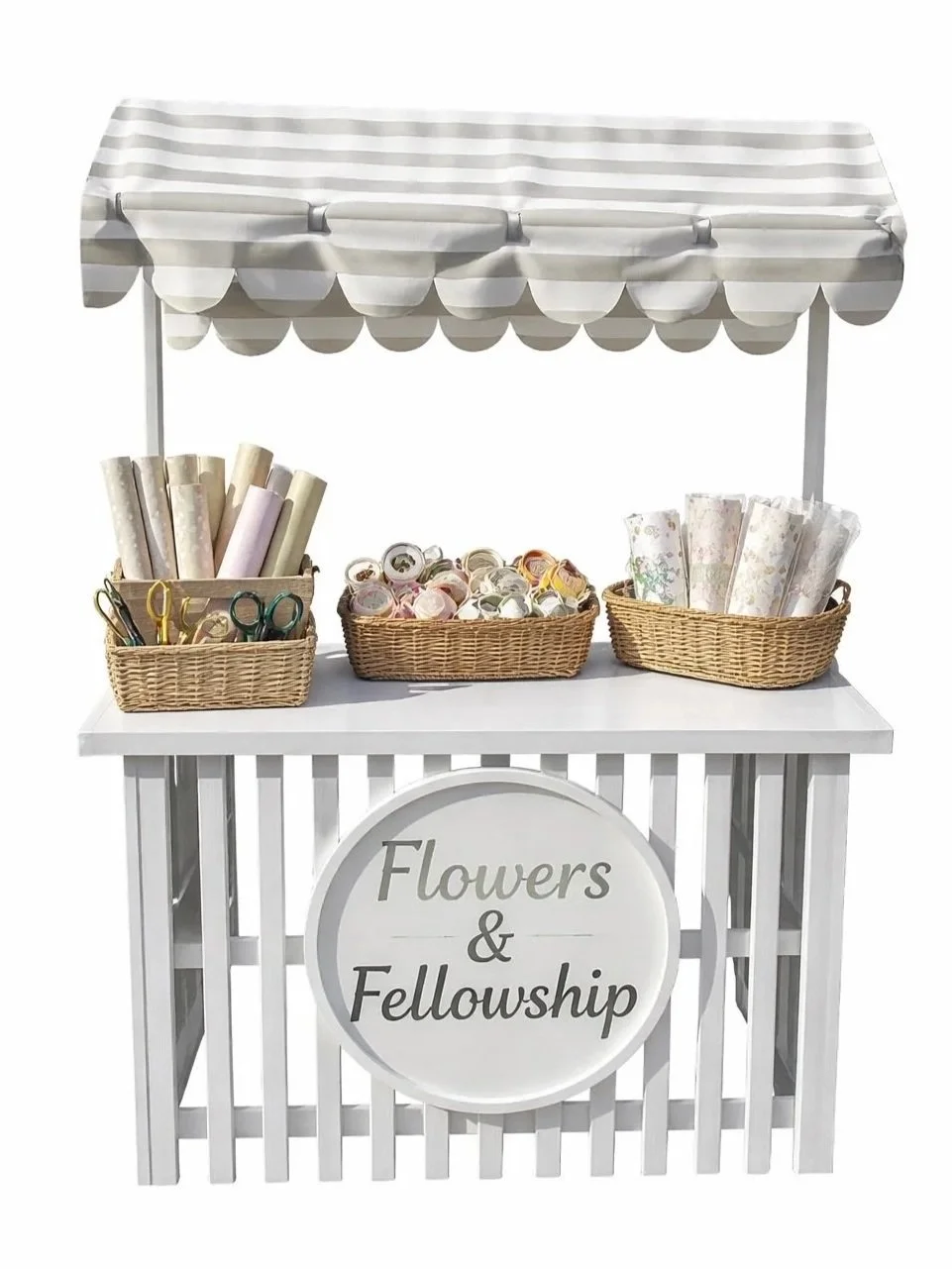 A white flower and fellowship booth with three wicker baskets filled with wrapping paper, scissors, and tape rolls, under a striped canopy with a sign reading 'Flowers & Fellowship' on the front.