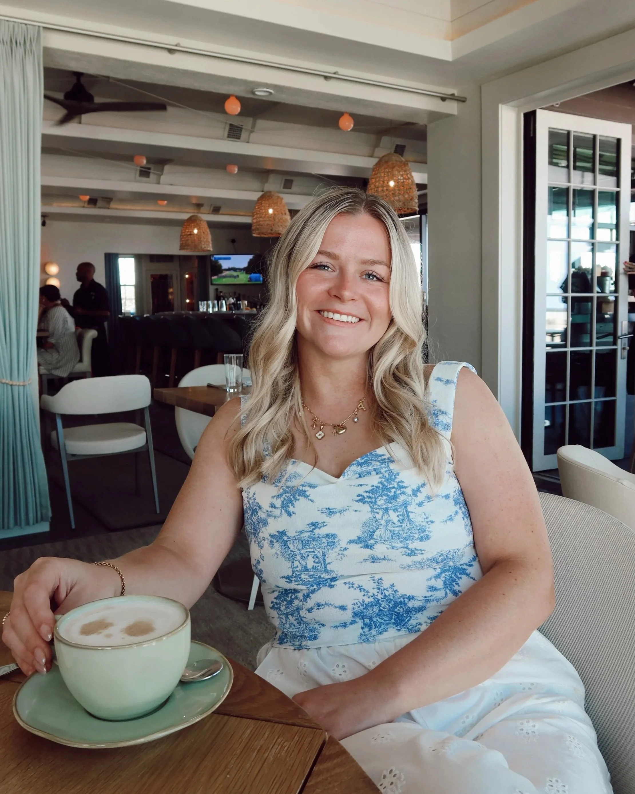 A woman with long blonde hair, wearing a blue and white dress with a scenic pattern, is sitting at a wooden table in a restaurant. She is smiling and holding a cup of coffee with foam topping. In the background, there are hanging lights, a television, and other restaurant patrons.