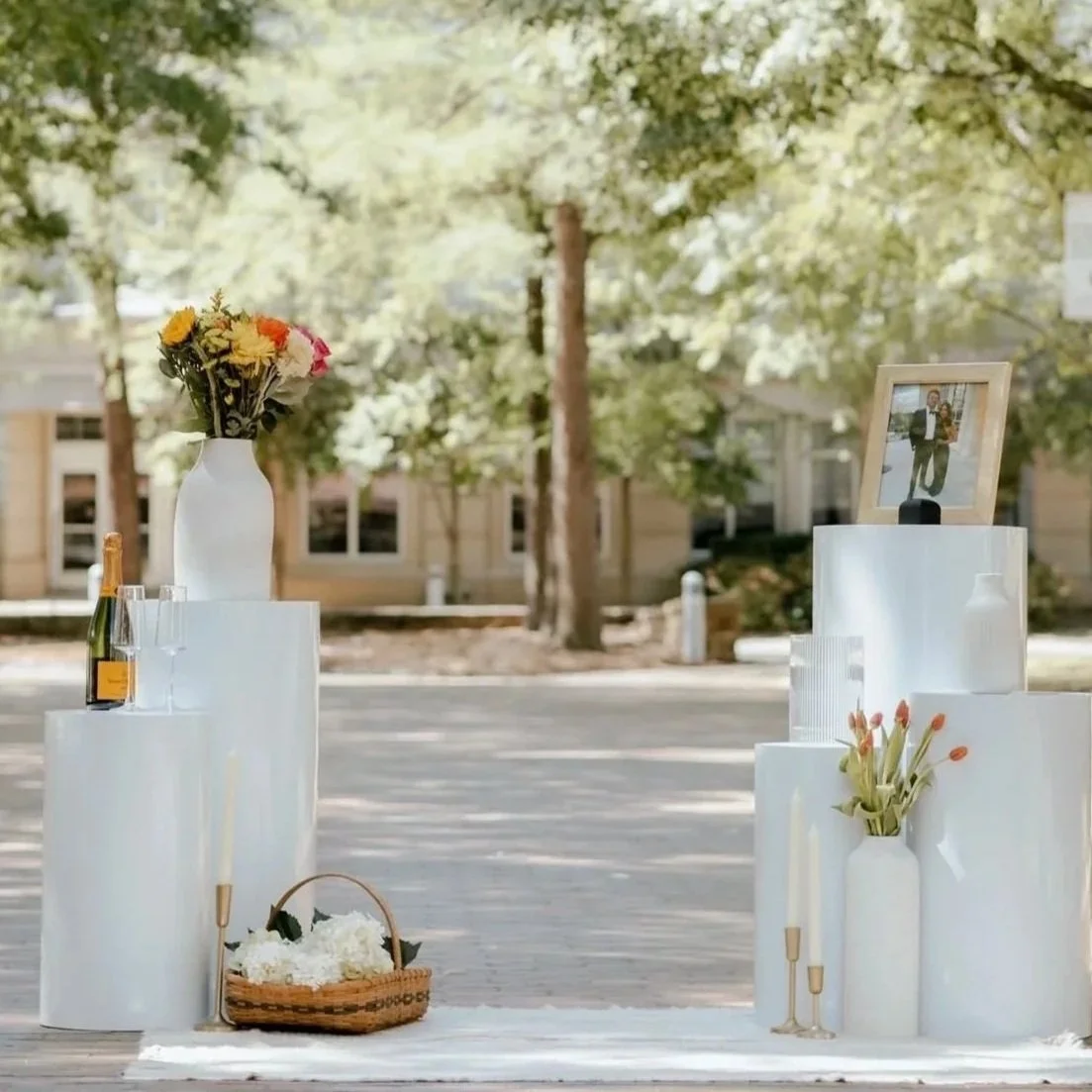 Arranged outdoor display with white pedestals holding a framed photograph, flowers in vases, a bottle of champagne with glasses, and a basket of white flowers, with trees and buildings in the background.