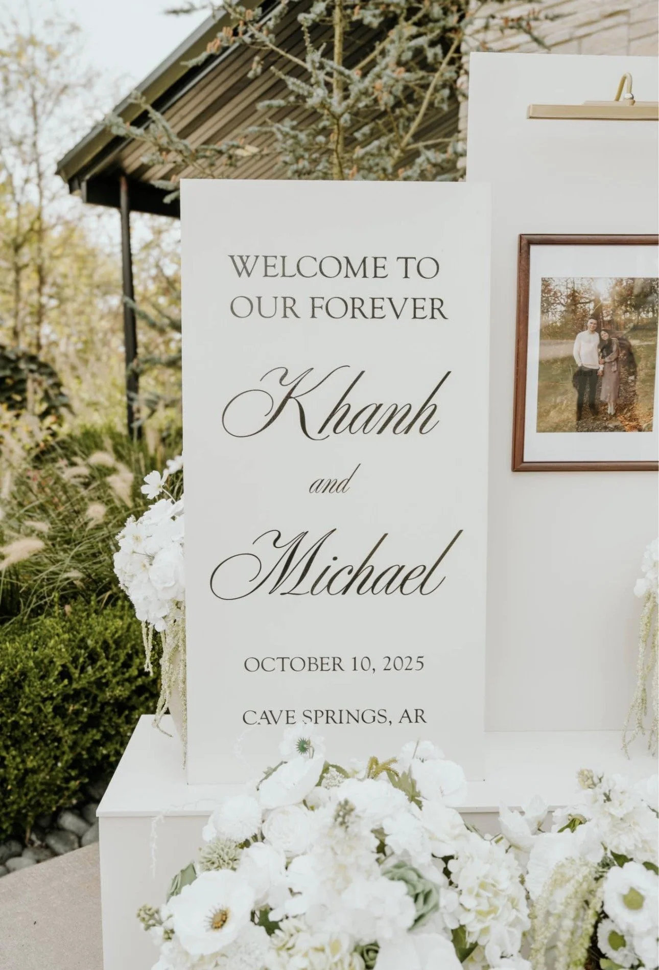 A wedding sign decorated with white flowers, displaying the names Khank and Michael, along with the date October 10, 2025, and location Cave Springs, AR. There is a framed photograph of a couple on the sign and a floral arrangement at the bottom.