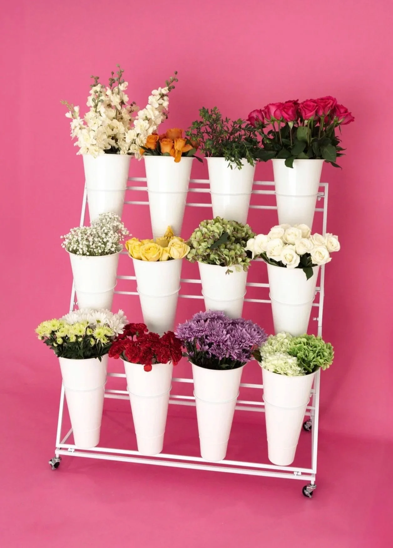Display of colorful flowers in white pots on a white metal stand against a pink background.