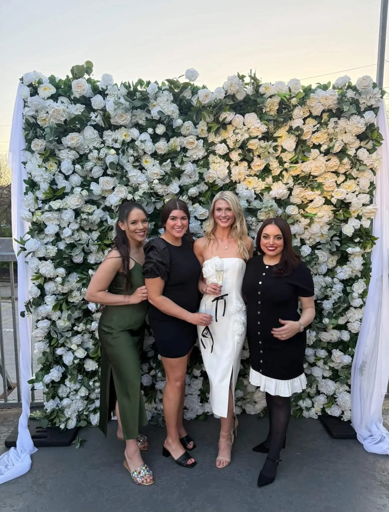 Four women in formal attire standing in front of a large floral backdrop made of white roses and green leaves, with one woman holding a glass of champagne.