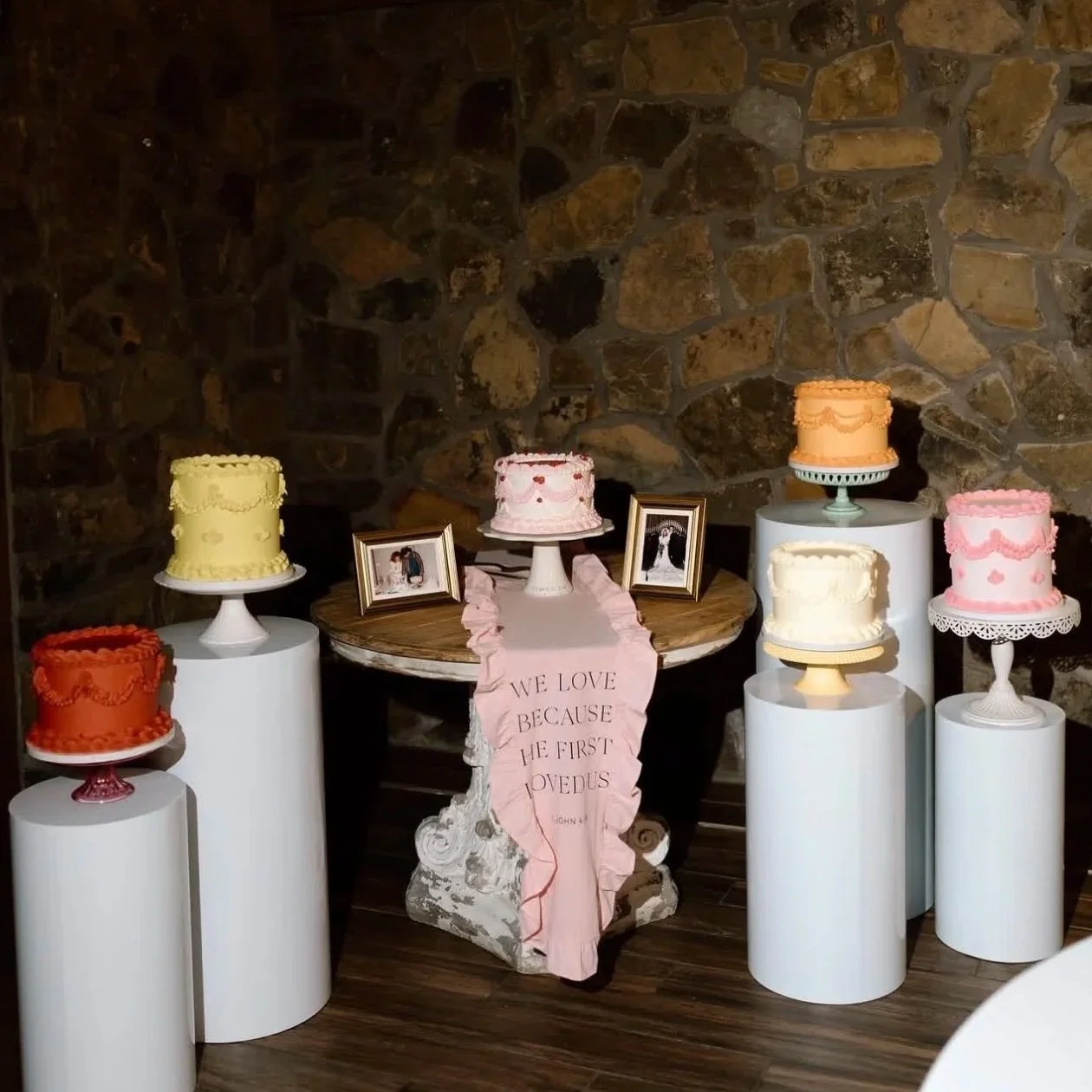 A display of six decorated cakes on white pedestals and stands arranged around a circular table against a stone wall background. The cakes are in orange, yellow, pink, white, and peach colors with decorative icing. Two framed pictures and a pink bann