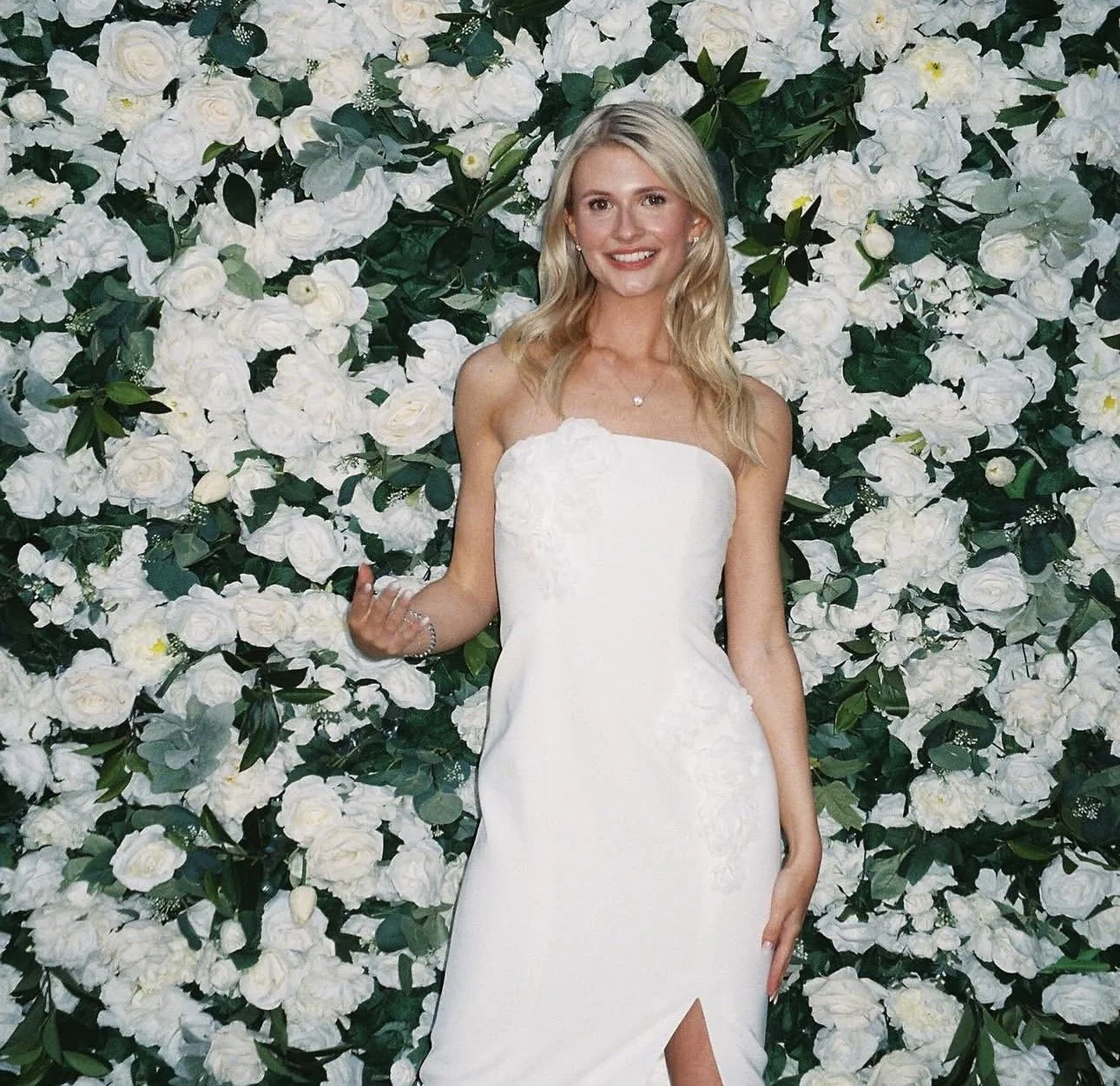 A woman in a white wedding dress standing in front of a floral wall of white roses and green leaves, smiling at the camera.