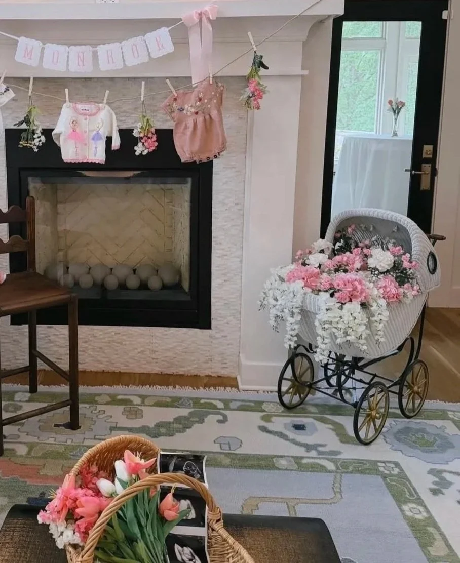 Decorated fireplace with a pink banner reading 'Monroe', stuffed animals and flowers hanging above, a vintage baby carriage filled with pink and white flowers, and baskets of flowers on a table.