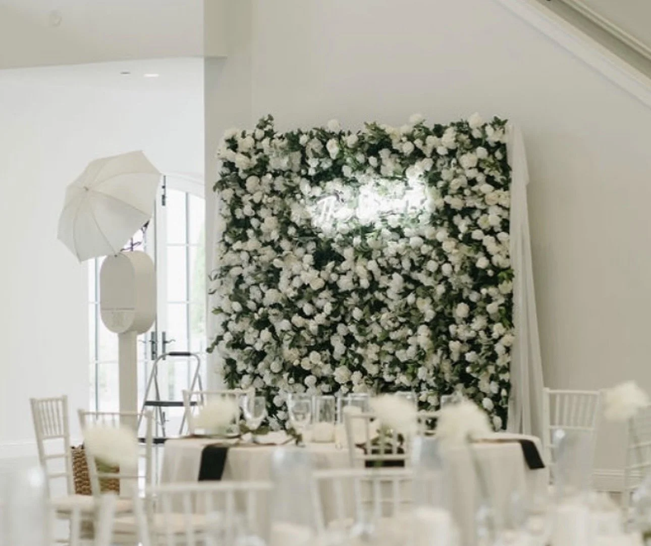 Wedding reception decorated with a large flower wall of white flowers, surrounded by white tables and chairs, and photo studio equipment like a flash, umbrella, and ladder in a bright room.