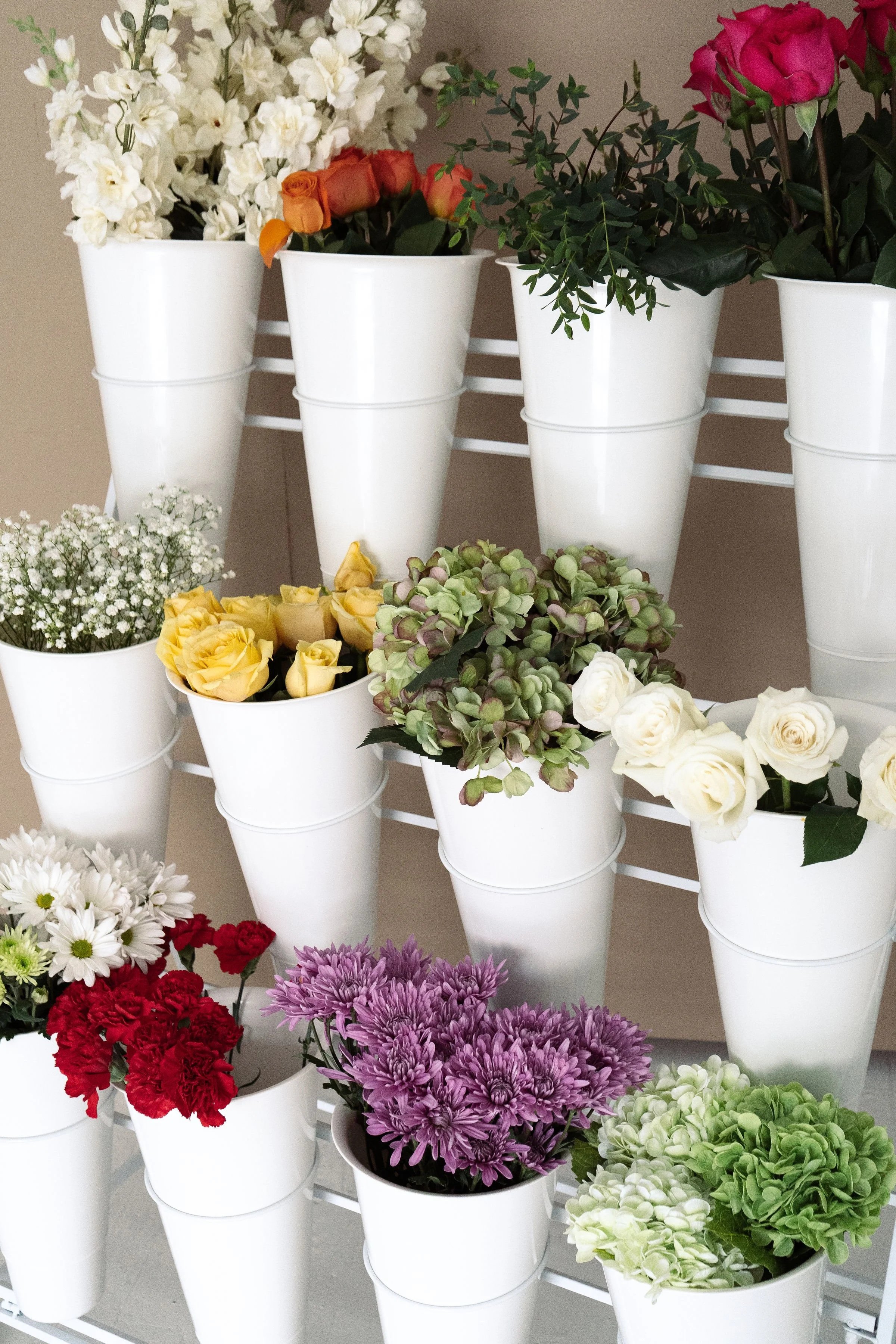Multiple white flower pots filled with various colorful flowers, including roses, daisies, and other blooms, arranged on a multi-tiered shelving unit.