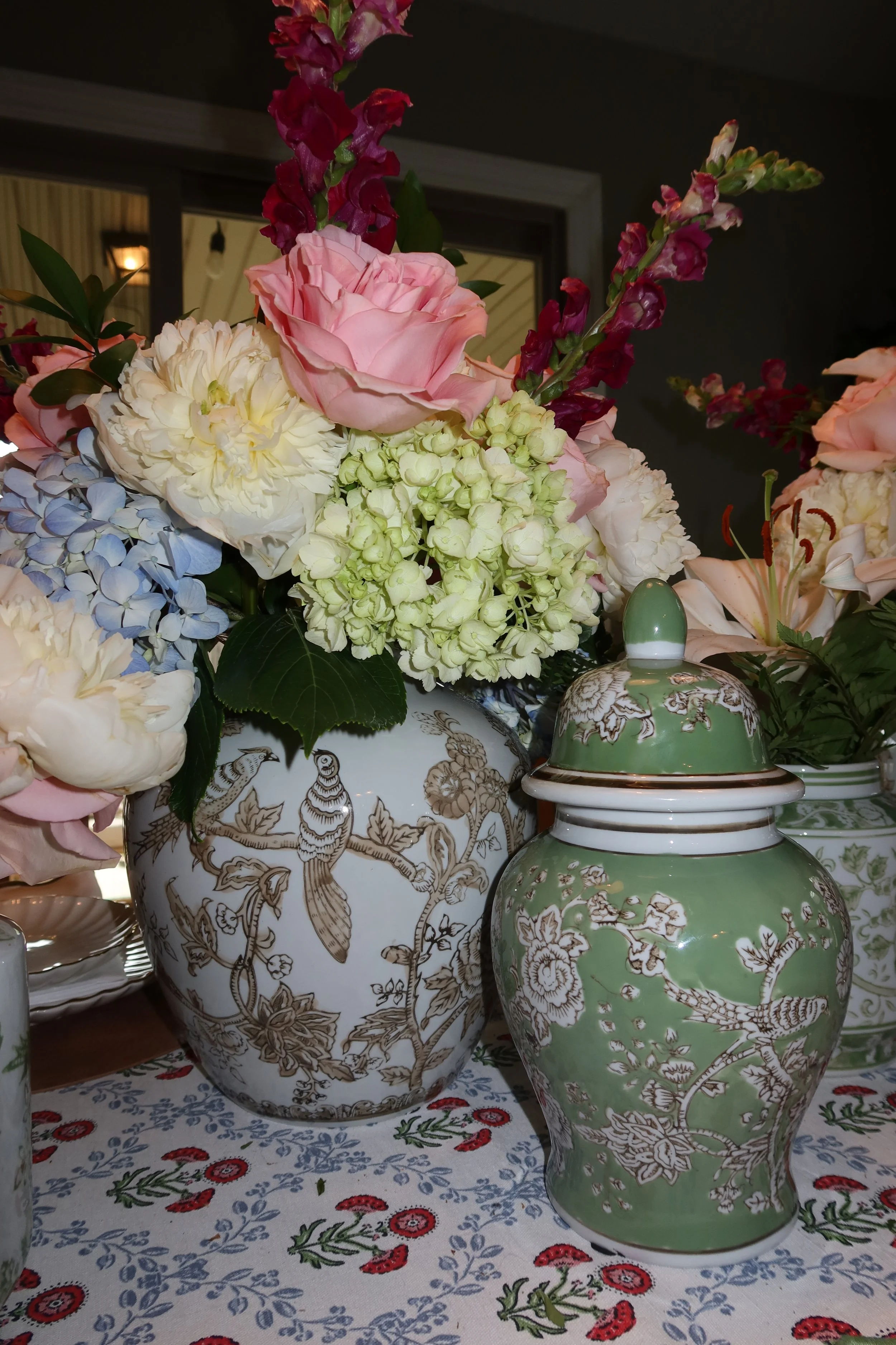 A floral arrangement featuring pink roses, white hydrangeas, and dark pink snapdragons in decorative ceramic vases with intricate gold patterns. The vases are placed on a table covered with a white cloth embroidered with red and blue floral motifs.