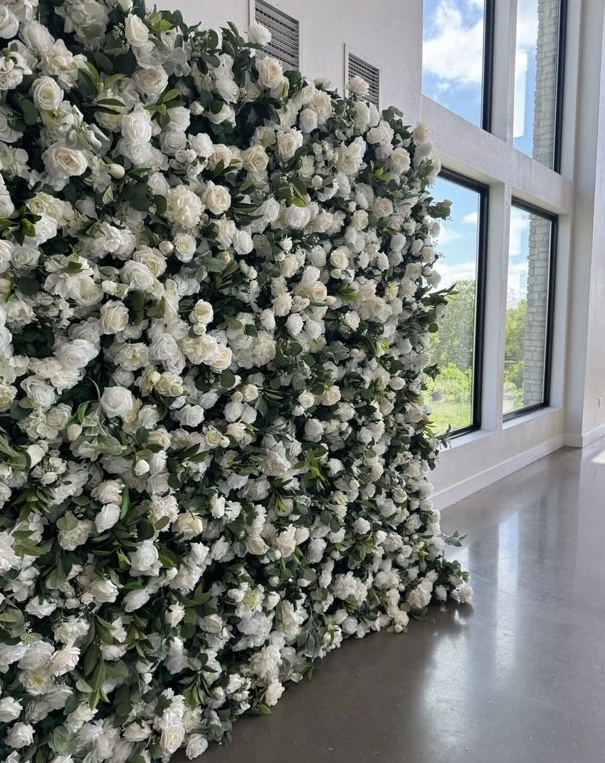 A large wall filled with white roses and green leaves, next to a glass window with a view of the outdoors.