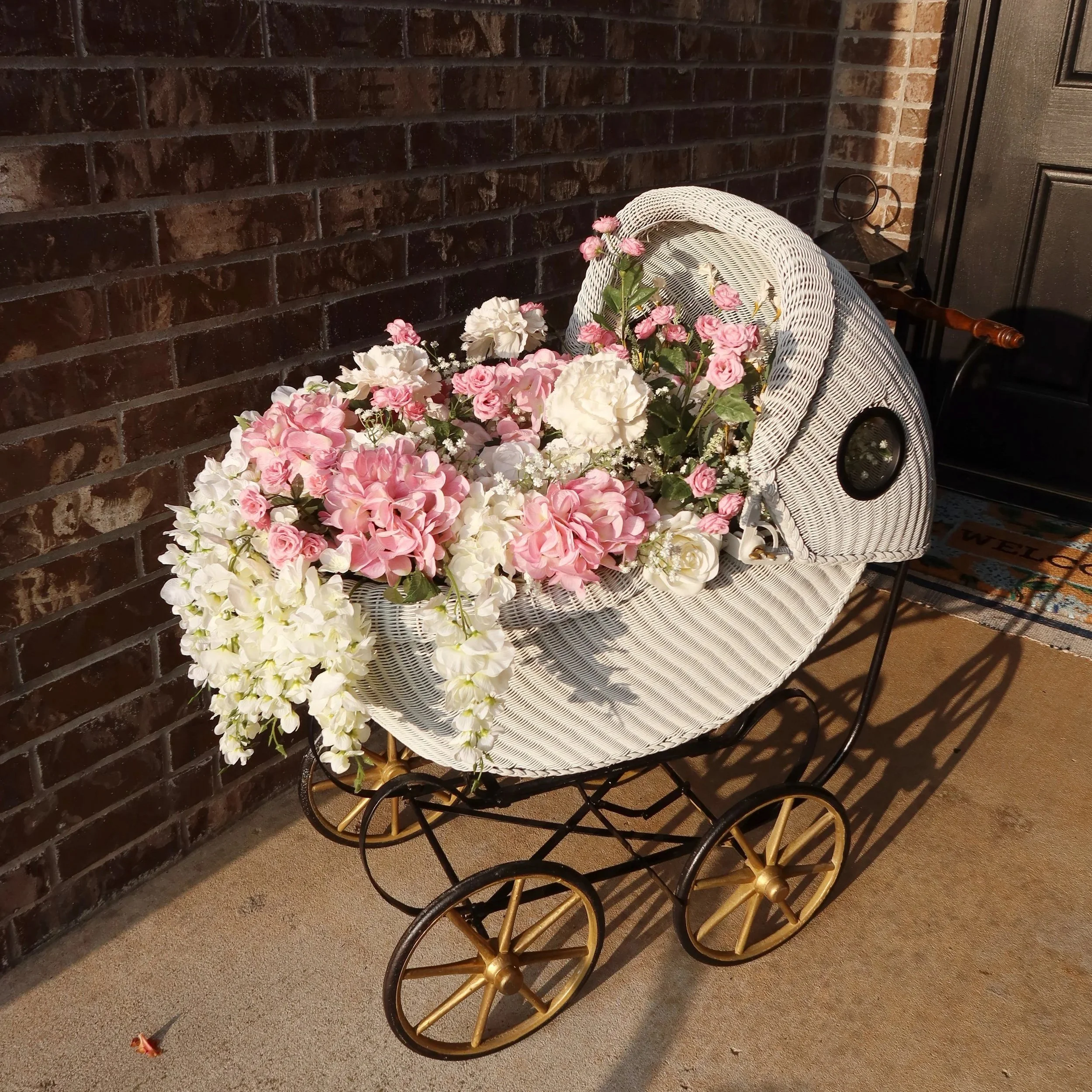 A white wicker carriage filled with pink and white flowers, placed outside against a brick wall.