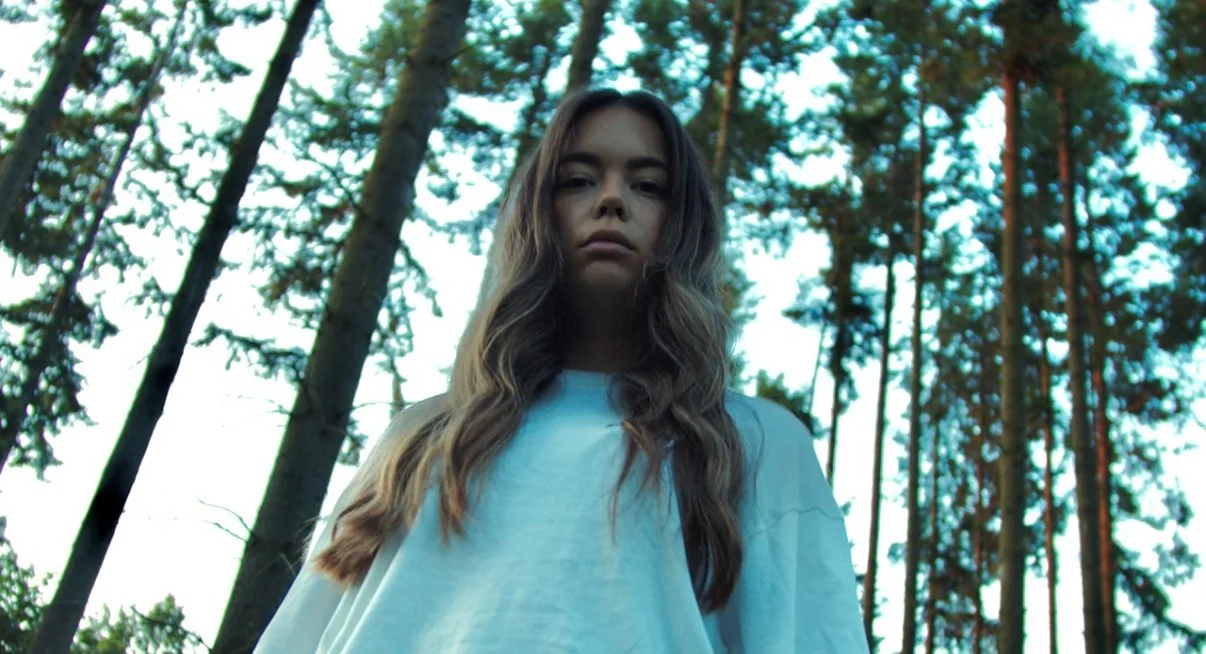 Junge Frau steht in einem Wald, blickt ernst und schaut nach unten.