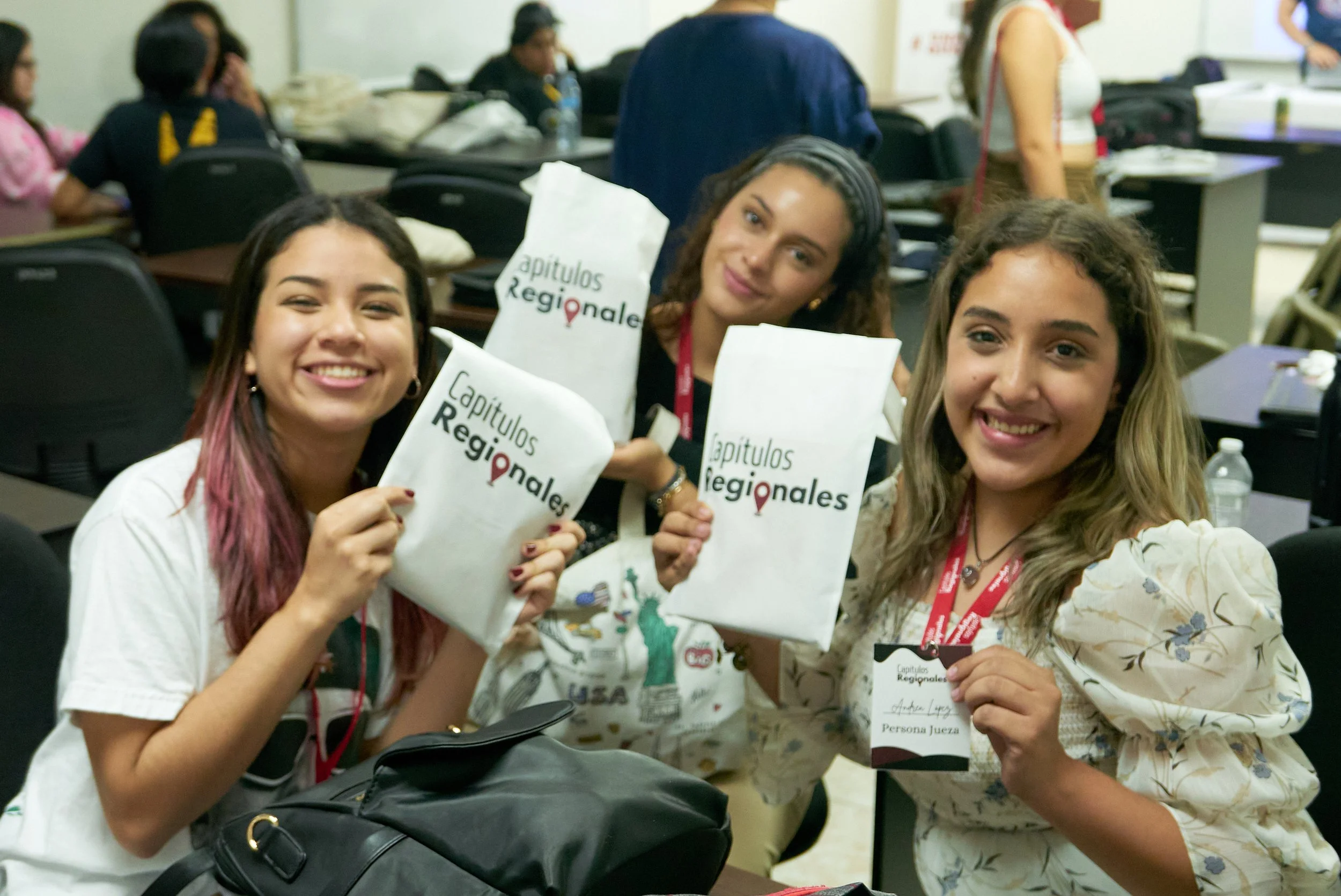 Tres jóvenes sonriendo y sosteniendo papeles con la leyenda "Capítulos Regionales" en un aula de conferencias, con varias personas de fondo en un entorno académico.