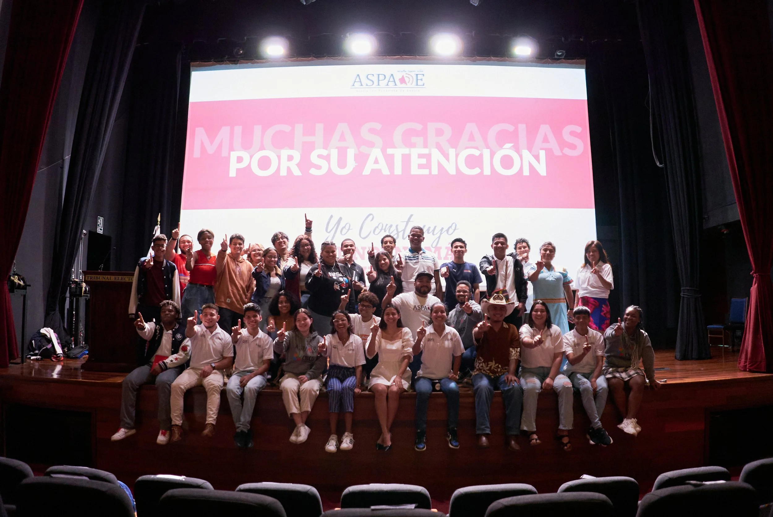 Grupo de personas en un escenario de teatro, algunos con vestido típico mexicano, celebrando y haciendo signo de número uno con la mano, en una presentación o evento. Fondo con pantalla que dice 'Muchas gracias por su atención' y el logo de ASPADE.