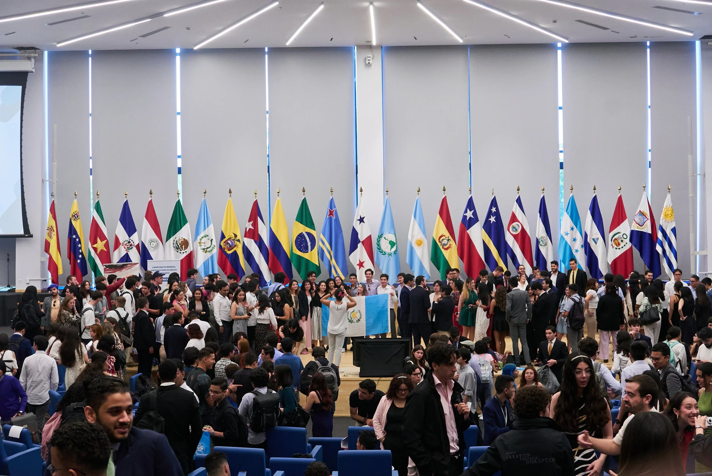 Gran sala con muchas personas sentadas y de pie, con banderas de varios países latinoamericanos en el fondo, en un evento o conferencia internacional.