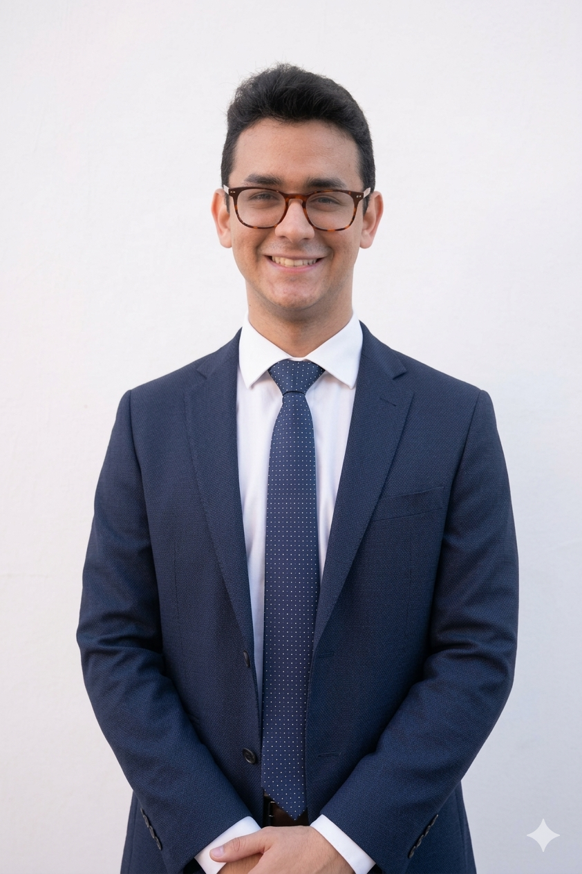Joven hombre con gafas, sonrisa, vestido con traje azul oscuro, camisa blanca y corbata de puntos, de pie frente a fondo blanco.