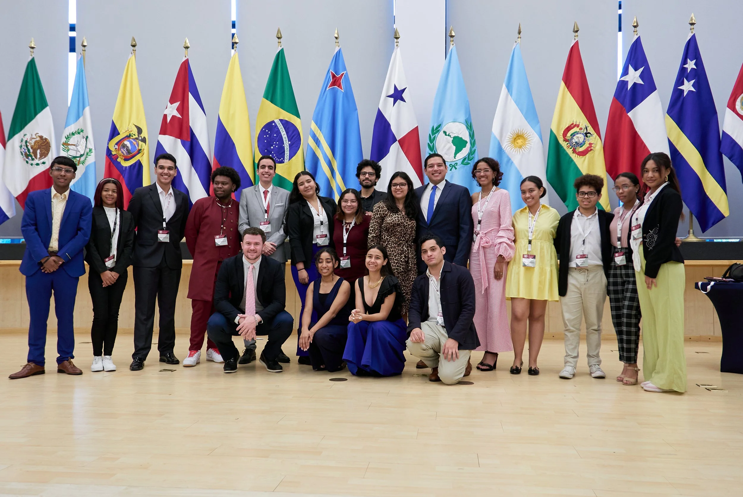 Grupo diverso de personas posando frente a banderas de países latinoamericanos en un evento formal.