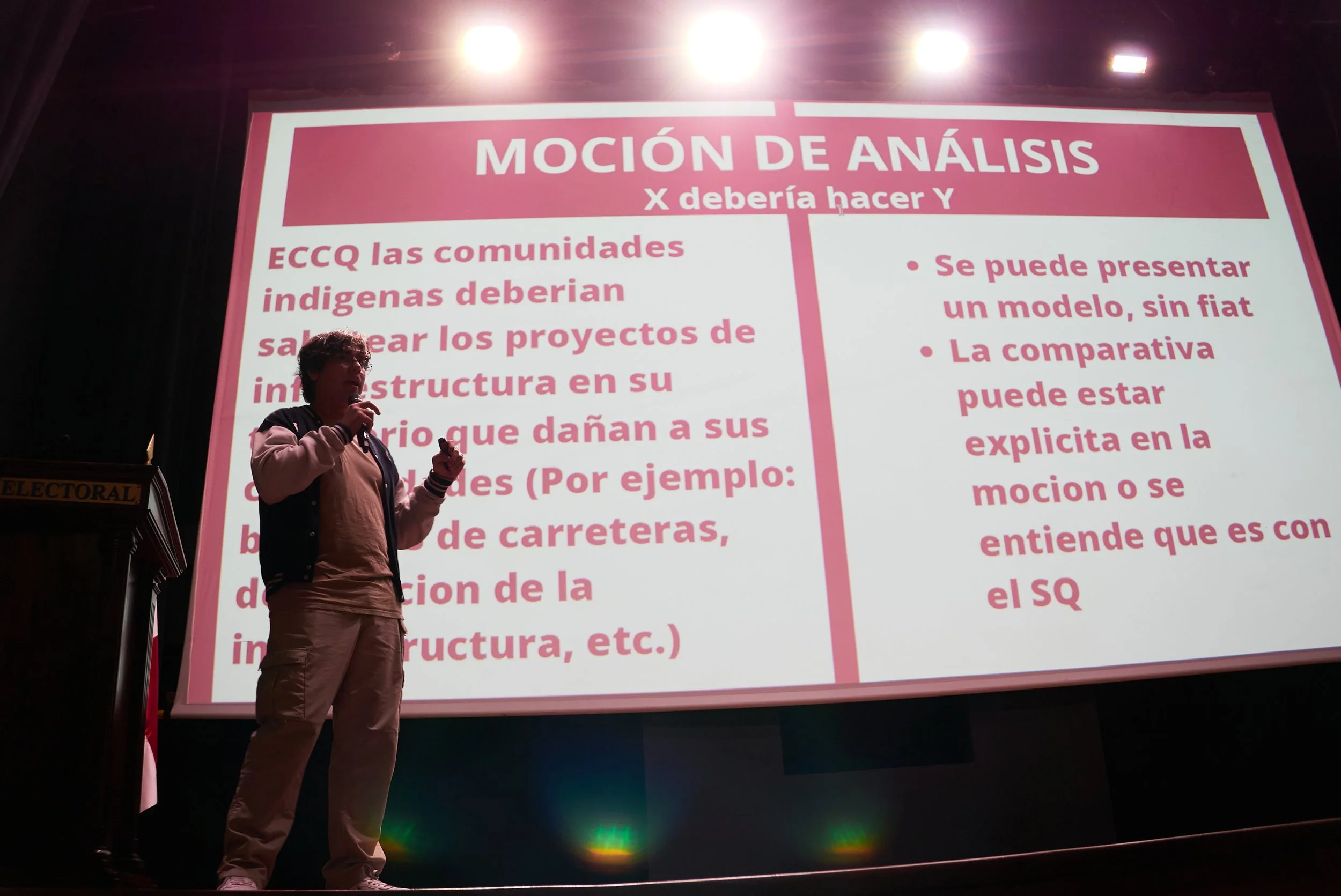 Persona hablando frente a una pantalla grande con una presentación en color rosa con texto en español. La pantalla muestra información sobre análisis y proyectos de infraestructura.