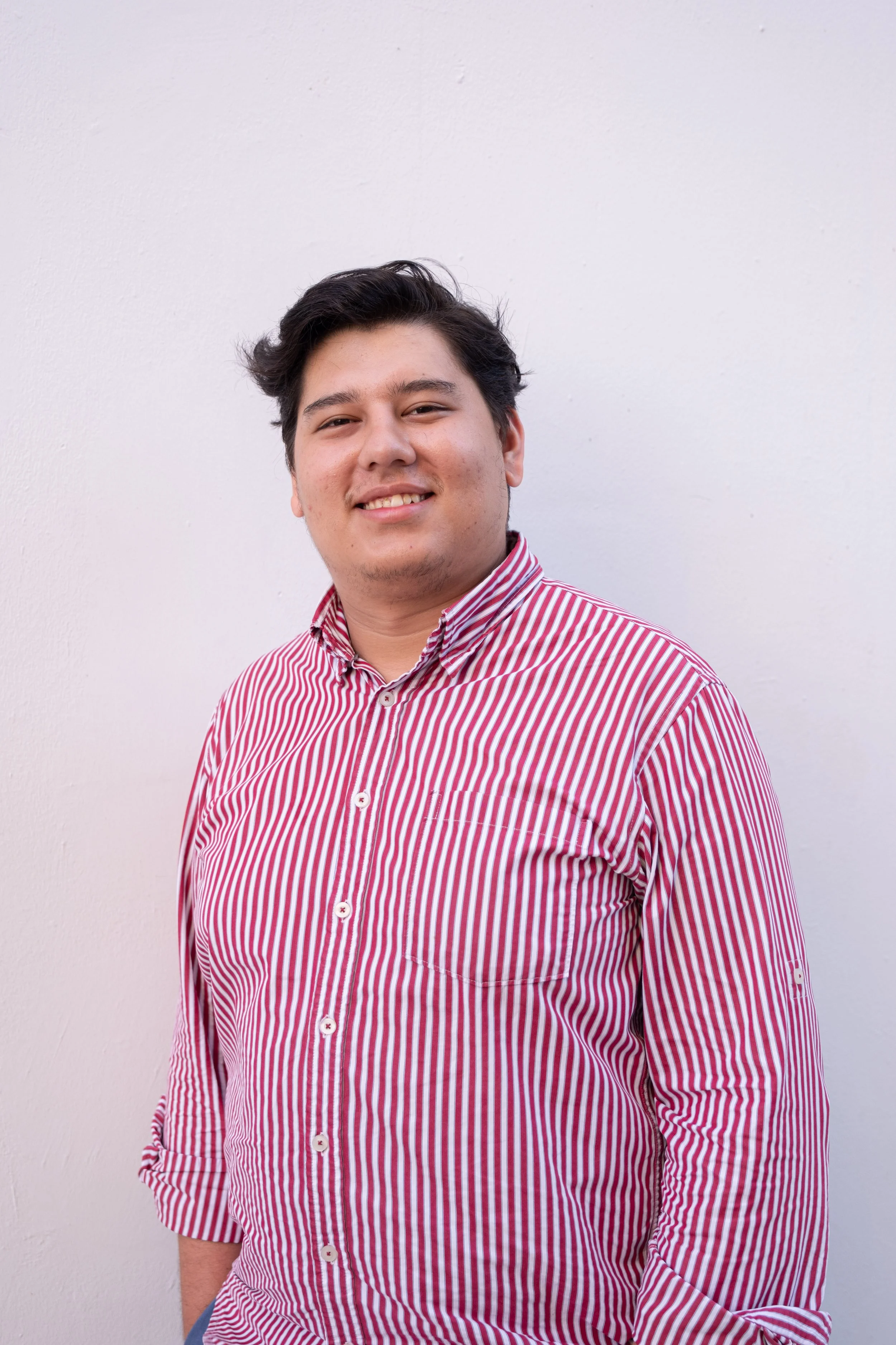 Joven hombre con cabello oscuro y piel clara, vistiendo una camisa de rayas rojo y blanco, sonriendo y posando frente a una pared blanca.