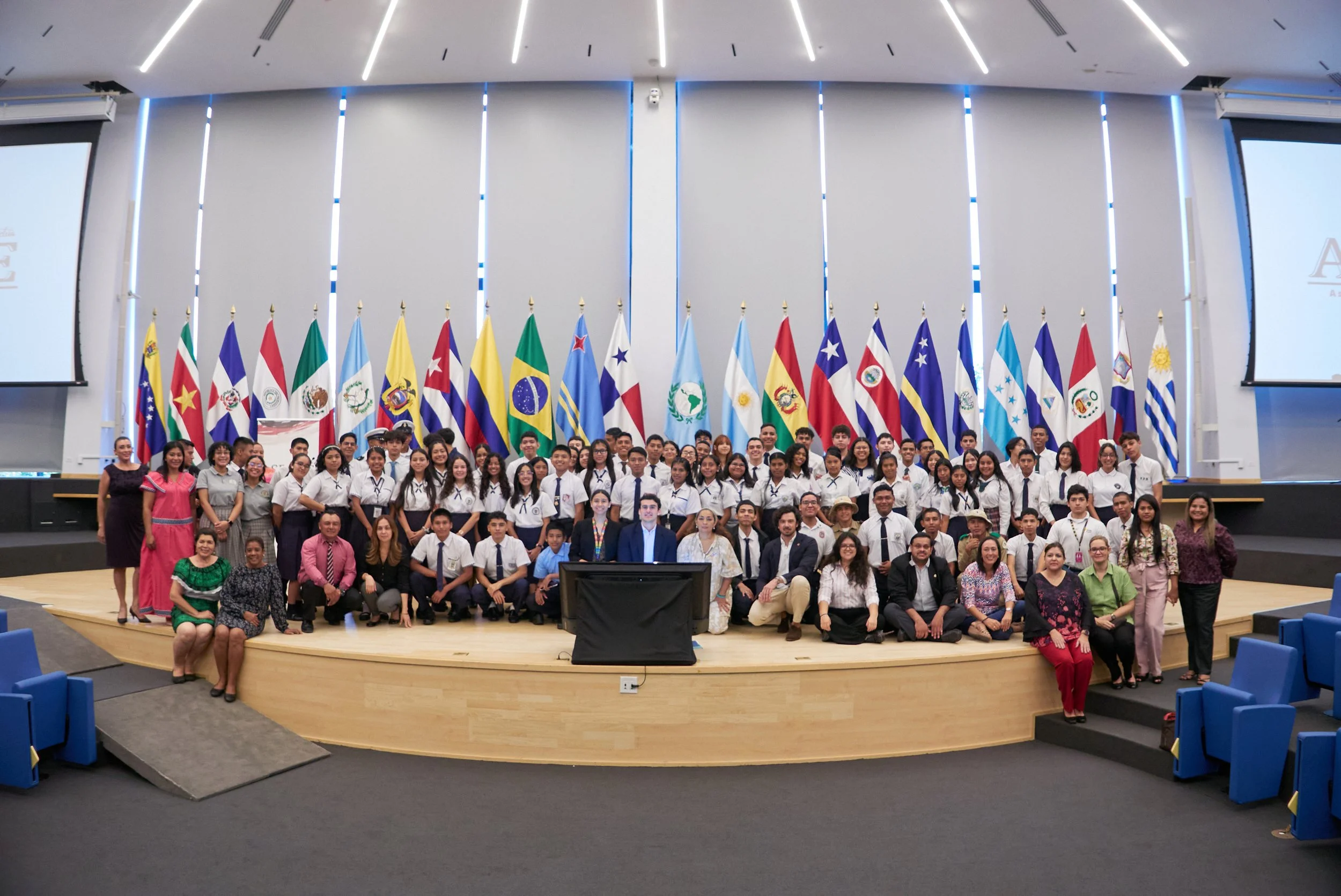 Grupo de personas en un auditorio con banderas de diferentes países en el fondo, probablemente en un evento internacional.