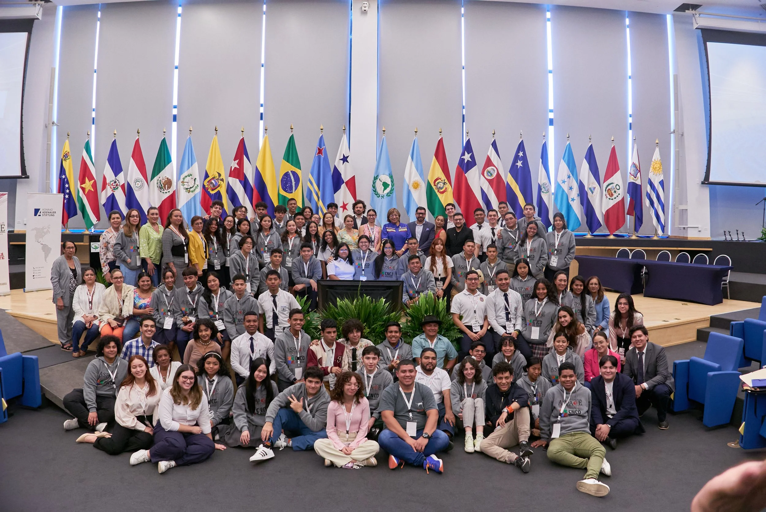 Grupo de jóvenes y adultos en un auditorio con banderas de países latinoamericanos al fondo. Todos posan para la foto, algunos con medallas y vestimenta casual o formal.